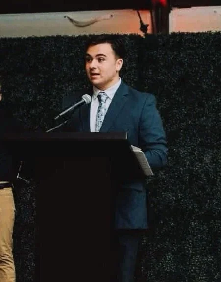 A young man in a suit speaking at a podium with a microphone, in front of a textured black backdrop.