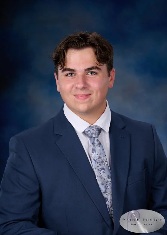 Young man in a navy suit, white shirt, and patterned tie posing against a dark blue background.