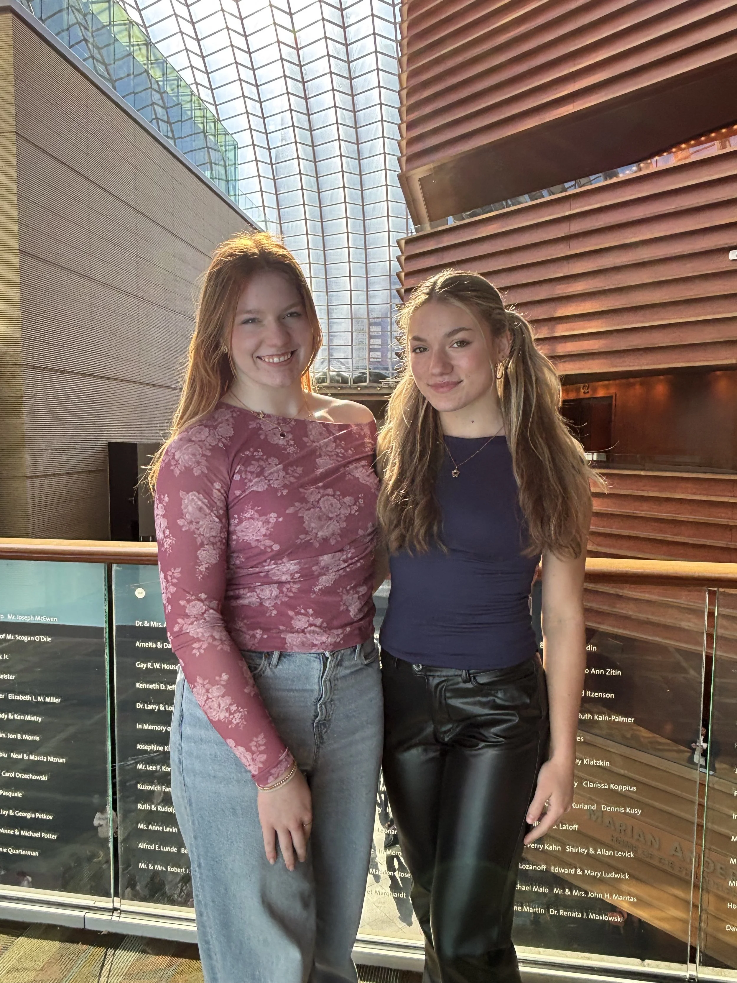 Two young women smiling and posing together indoors near a glass wall with modern architecture in the background.
