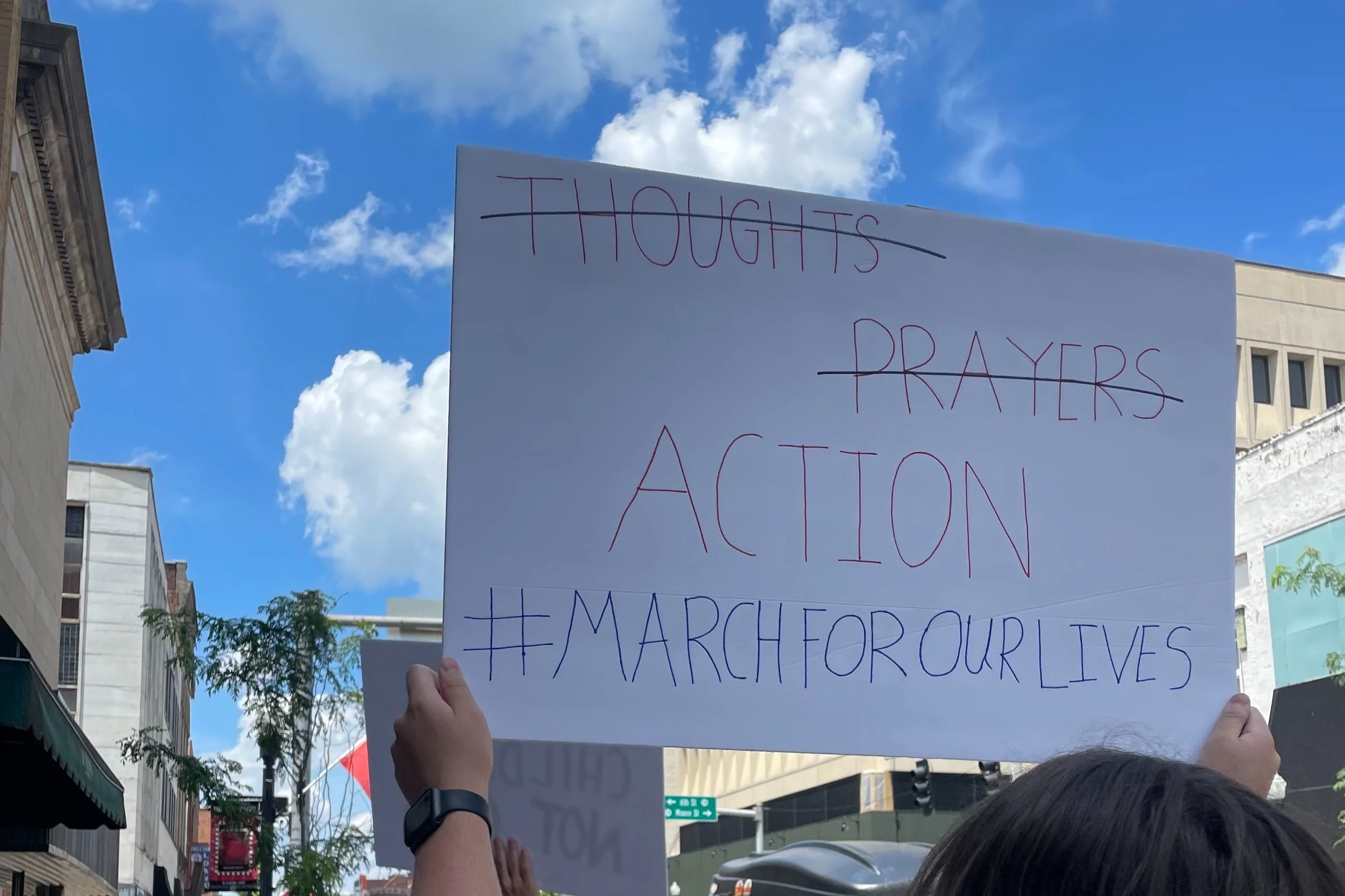 A protestor is holding a white sign with handwritten words in red and purple banners that reads, "THOUGHTS PRAYERS ACTION #MARCHFOROURLIVES". The photo was taken from behind the protestor during the day with blue sky and clouds in the background. Buildings and trees are also visible.