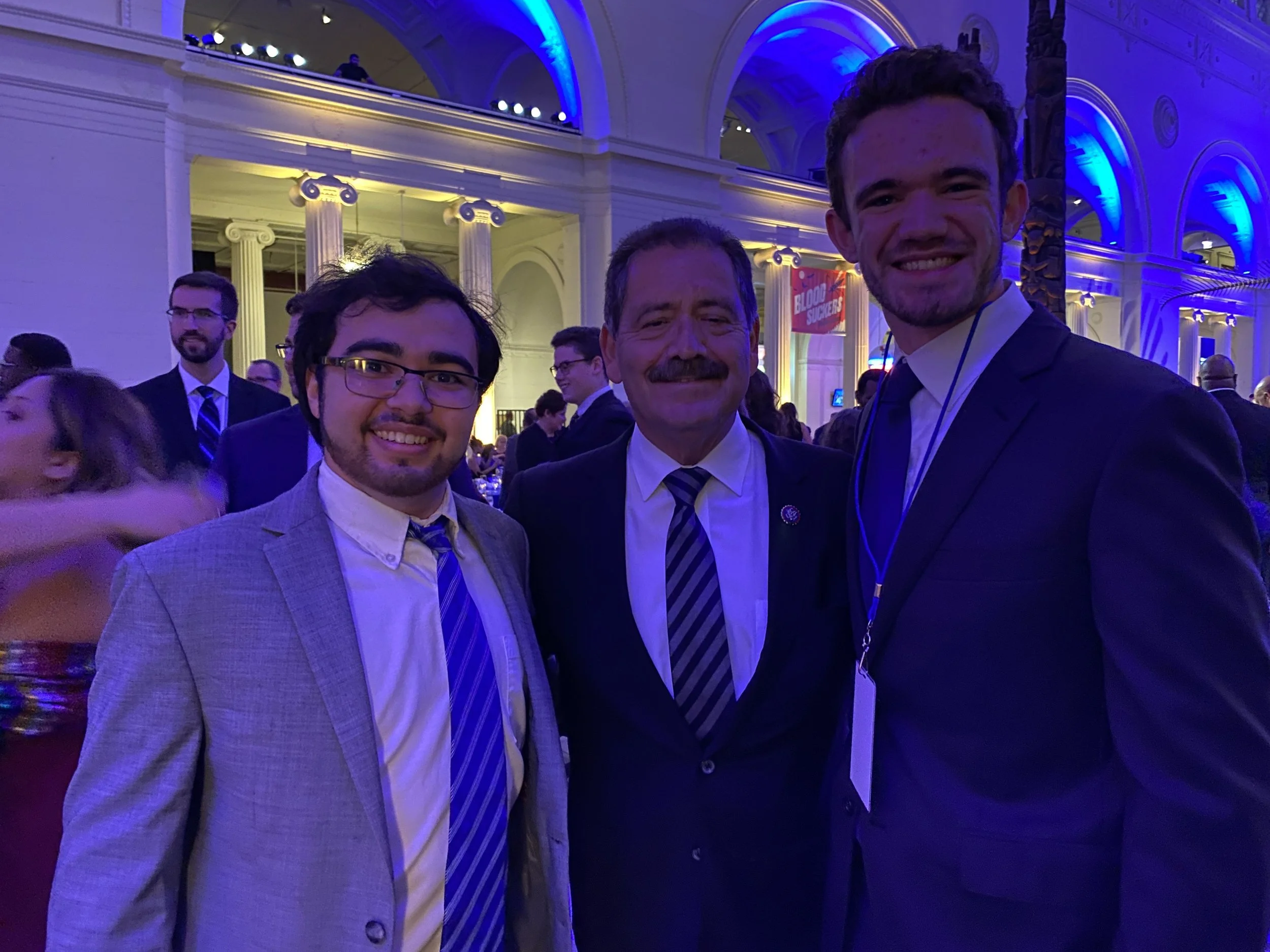 Three men dressed in suits posing for a photo at an indoor event with architecture featuring tall columns, arches, and vibrant purple and yellow lighting.