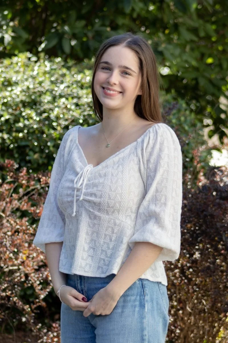A young woman with long brown hair and a big smile stands outdoors in front of green bushes and trees, wearing a white textured blouse with a drawstring at the neckline and blue jeans.