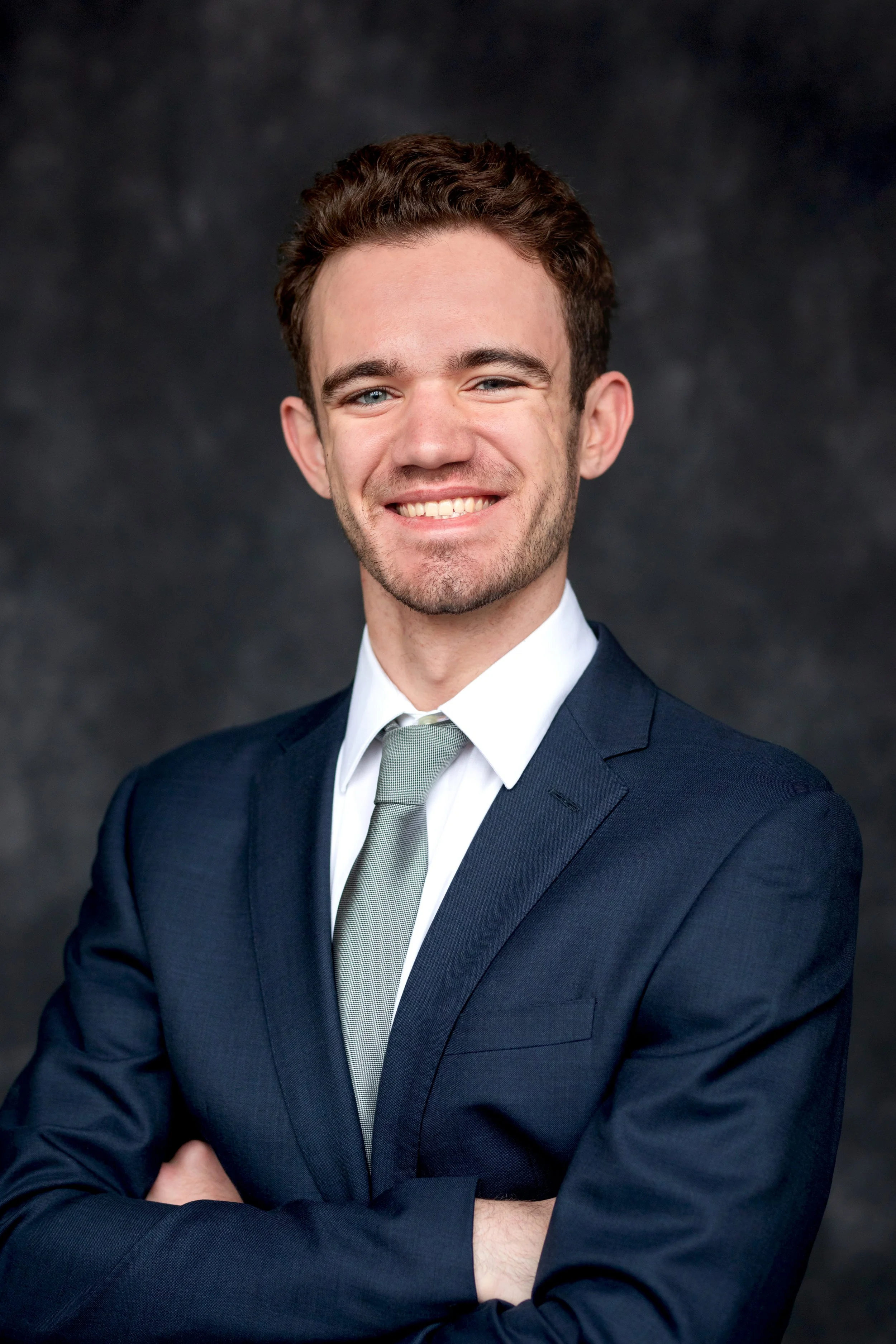 A man in a navy blue suit, white shirt, and light gray tie posing with arms crossed in front of a dark background.