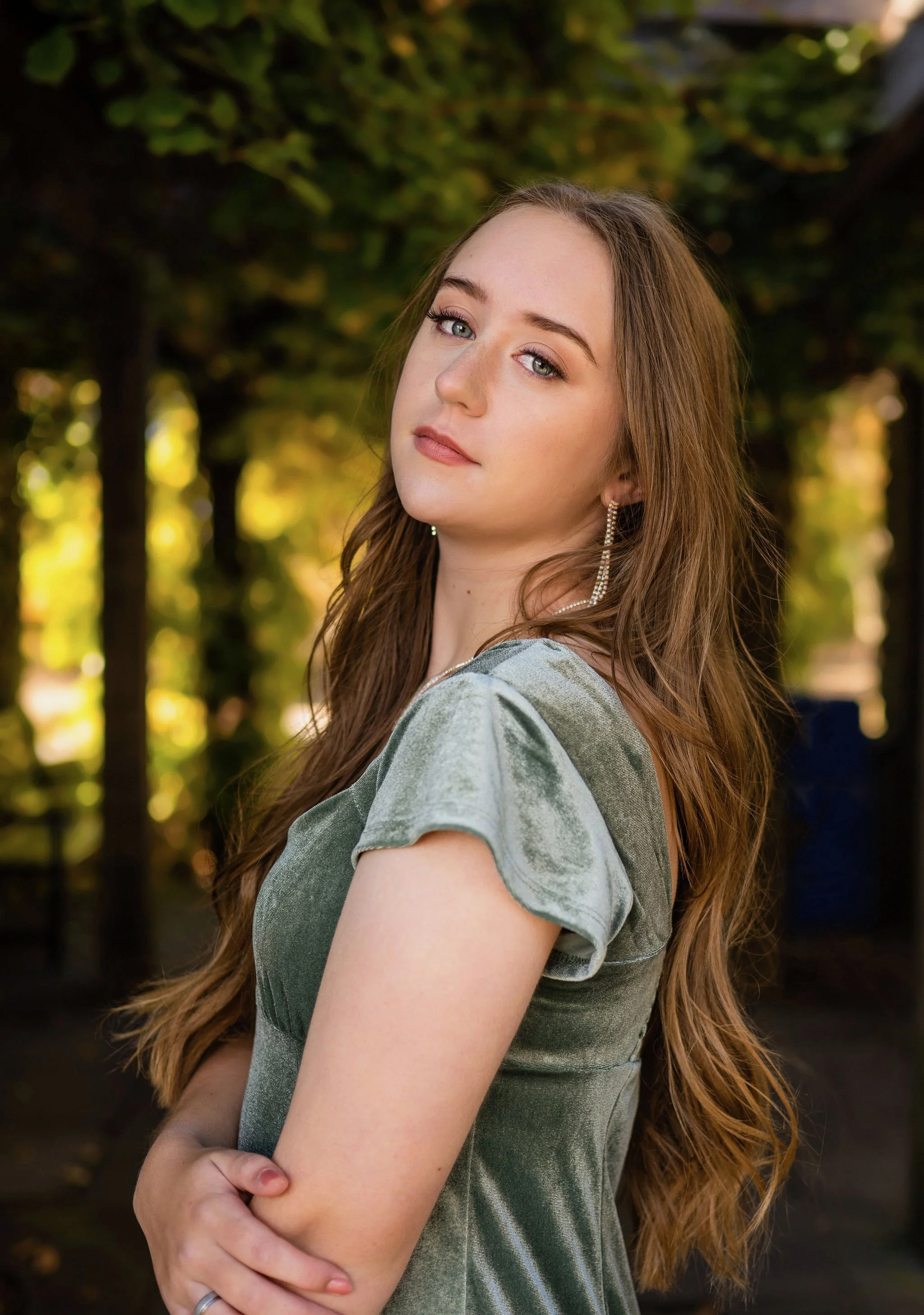 A young woman with long, wavy red hair, blue eyes, and a light skin tone looking at the camera outdoors. She is wearing silver earrings and a velvet dress with puffy sleeves. The background features blurred trees with green and yellow leaves, indicating an autumn setting.