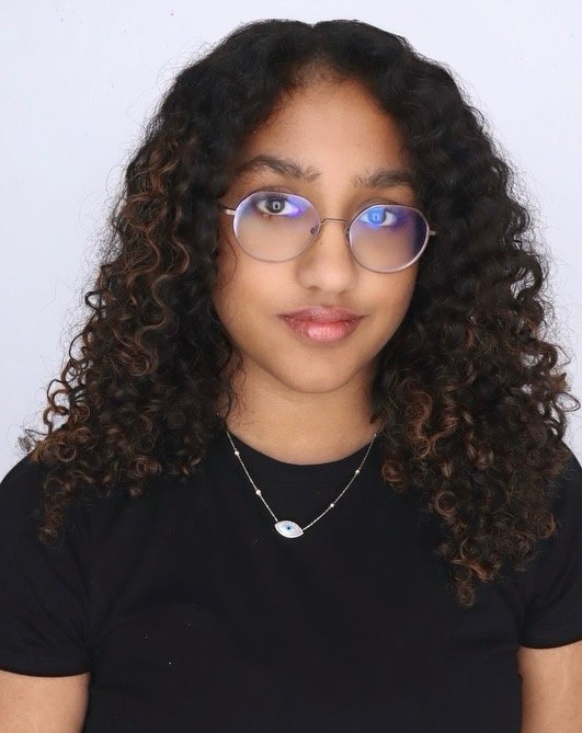 A young woman with curly dark hair, wearing glasses, a black shirt, and a gold necklace with an eye charm, smiling at the camera against a plain white background.