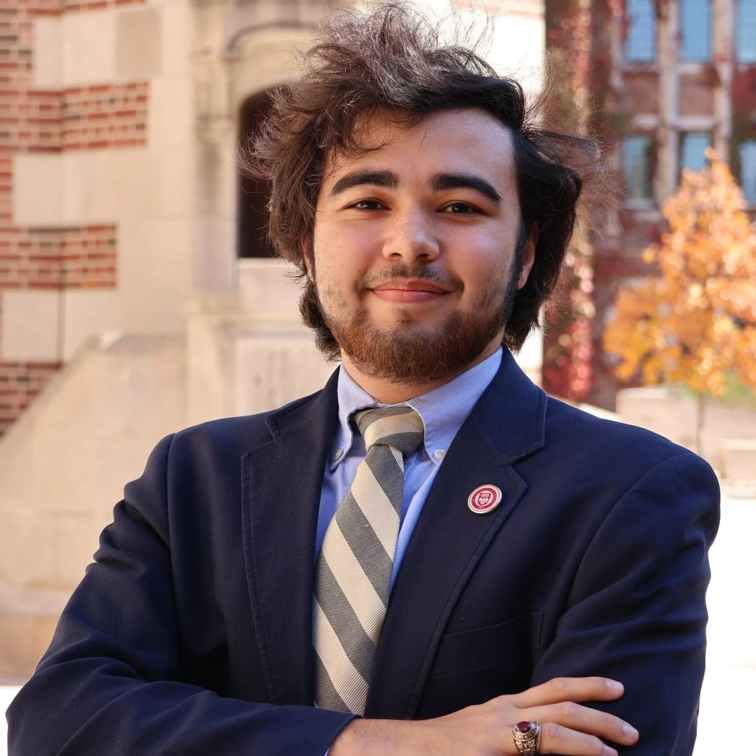 A young man with dark, messy hair and a beard wearing a navy blue suit, white shirt, and striped tie, standing outdoors with autumn foliage in the background.