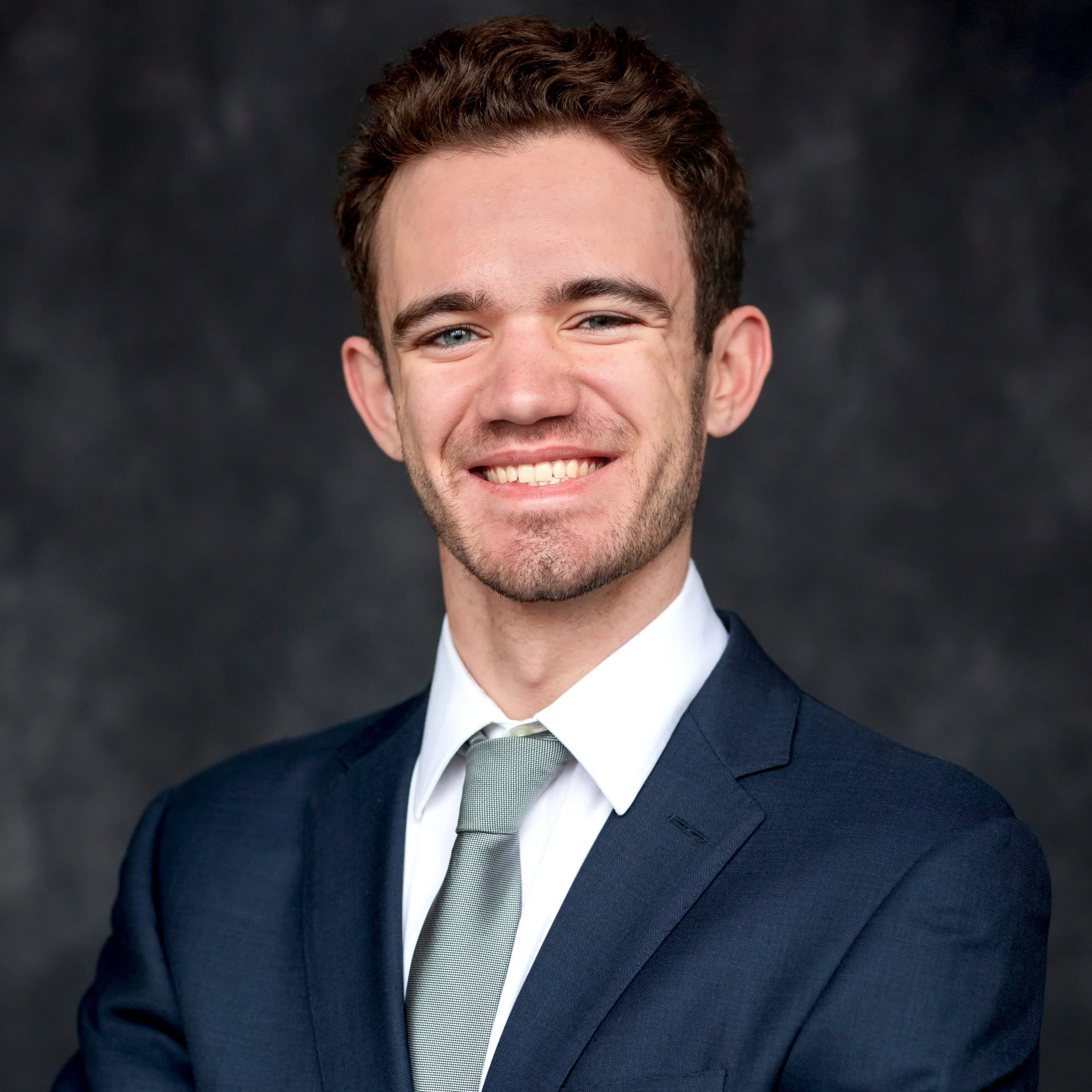 Close-up of a young man with brown curly hair, blue eyes, smiling, wearing a dark suit, white shirt, and gray tie, against a black background.