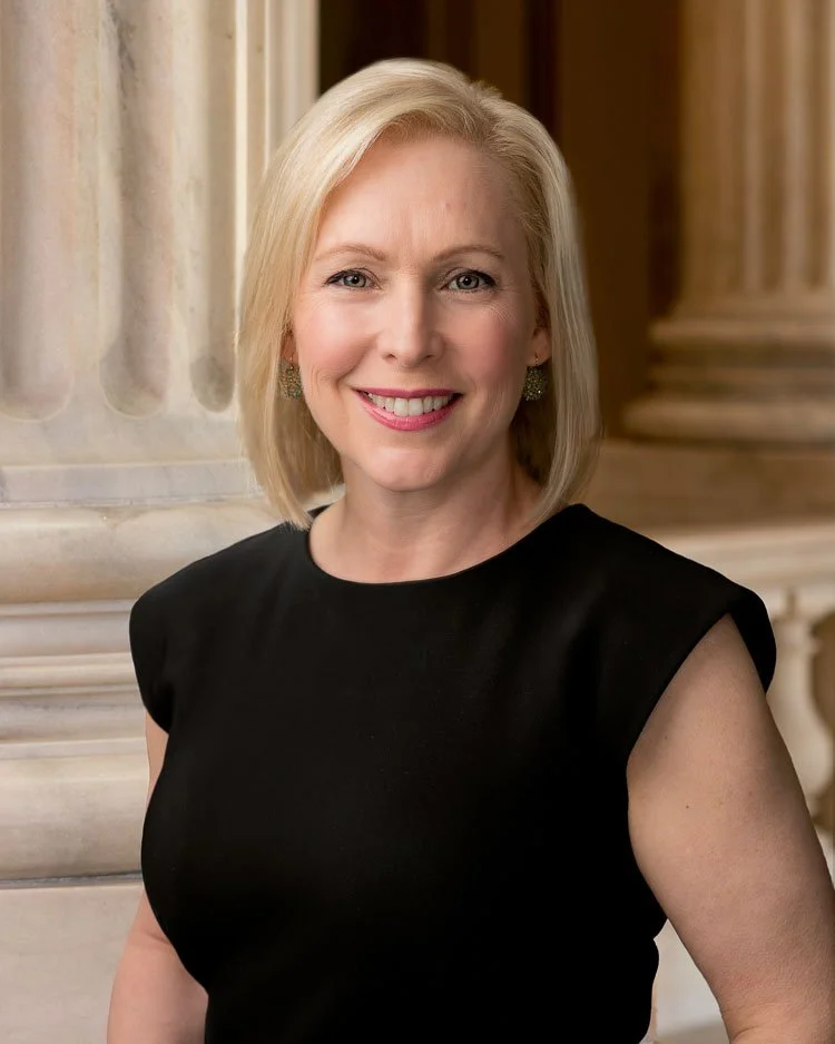 A smiling blonde woman wearing a black sleeveless top standing in front of a marble columned background.