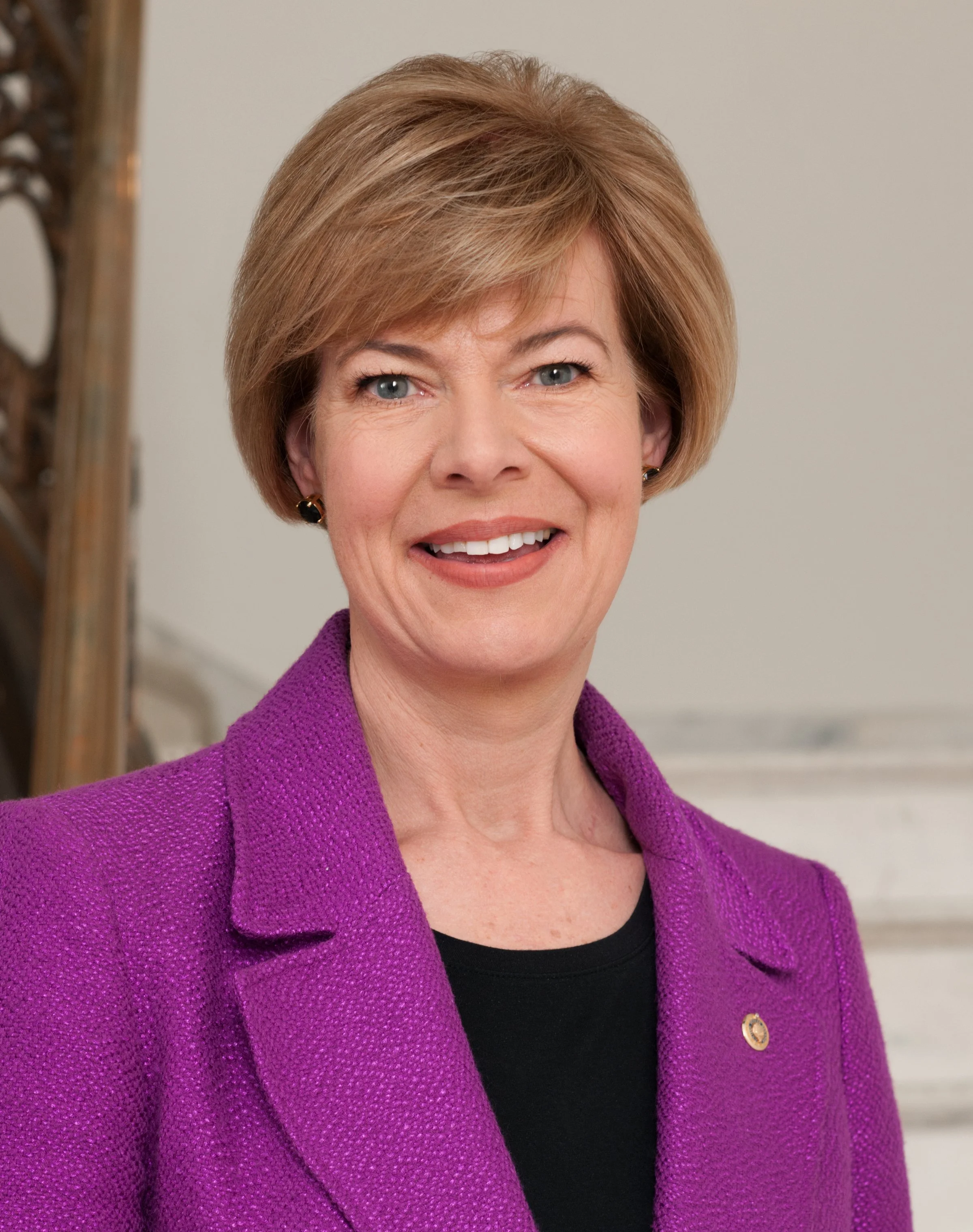 Close-up of a woman with short, light brown hair and blue eyes smiling, wearing a purple blazer with a circular pin and a black top.