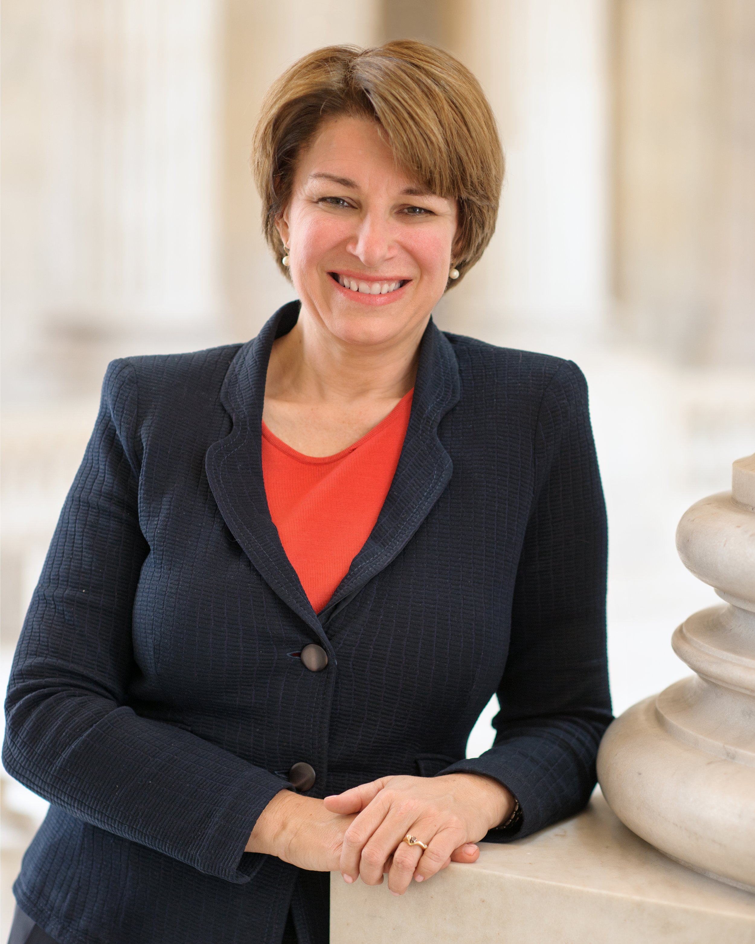 A woman with short brown hair, wearing a navy blazer and a coral top, smiling and leaning on a marble counter with a decorative sculpture beside her.