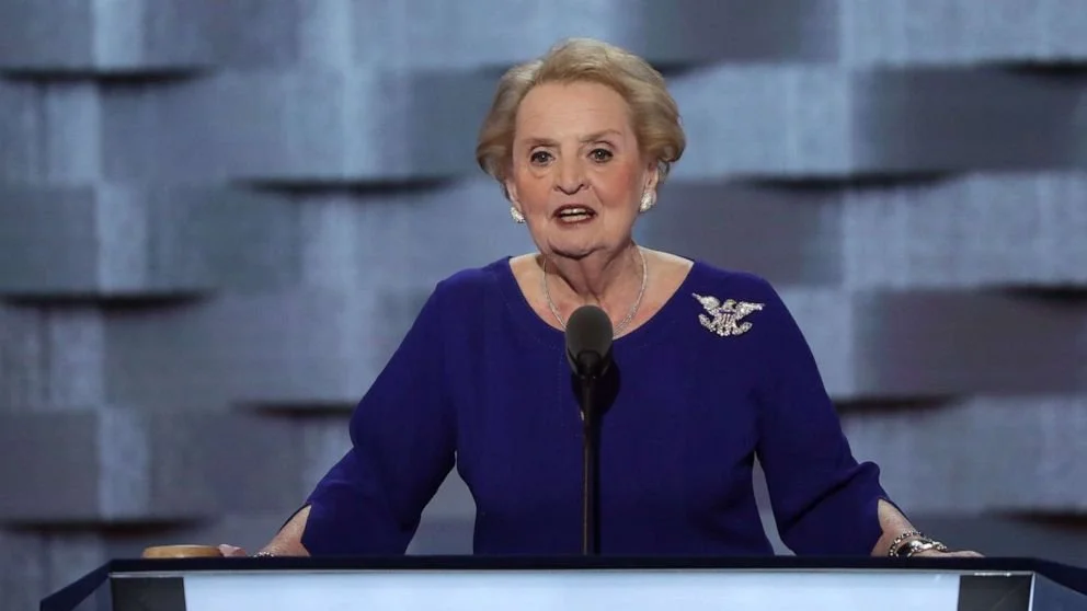 An elderly woman with short, curly blonde hair speaking at a podium with a microphone. She is wearing a dark blue dress with a decorative brooch and earrings.