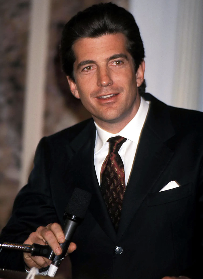 A man in a dark suit holding a microphone, with brown hair and a white shirt, appears to be speaking at an event.
