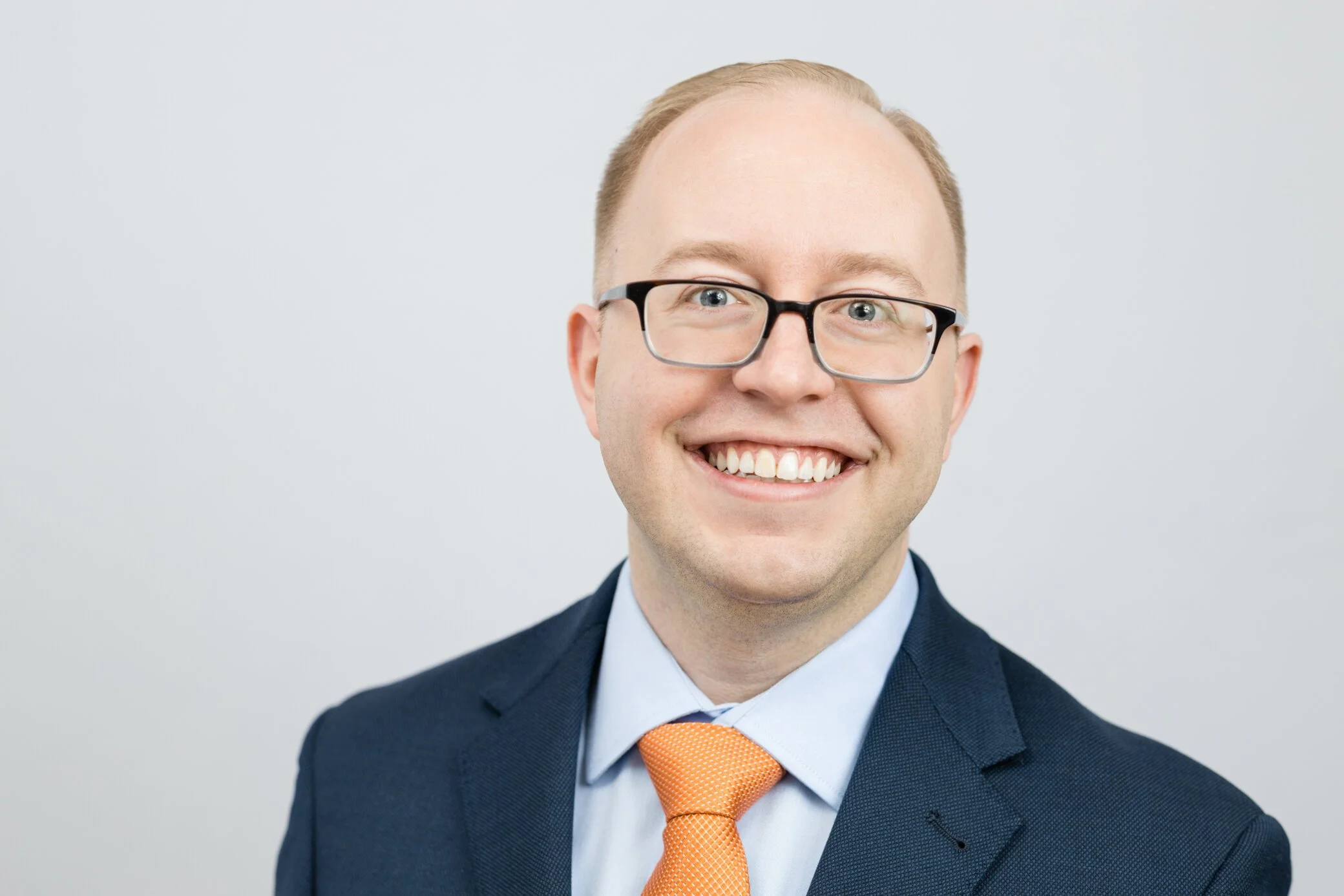 Close-up of a smiling young man with glasses, dressed in a navy blue suit, light blue shirt, and orange tie, against a plain white background.