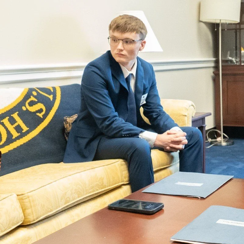 Young man with glasses in a blue suit sitting on a yellow sofa, in a room with a lamp and wooden furniture, with a smartphone and folders on a table in front of him.