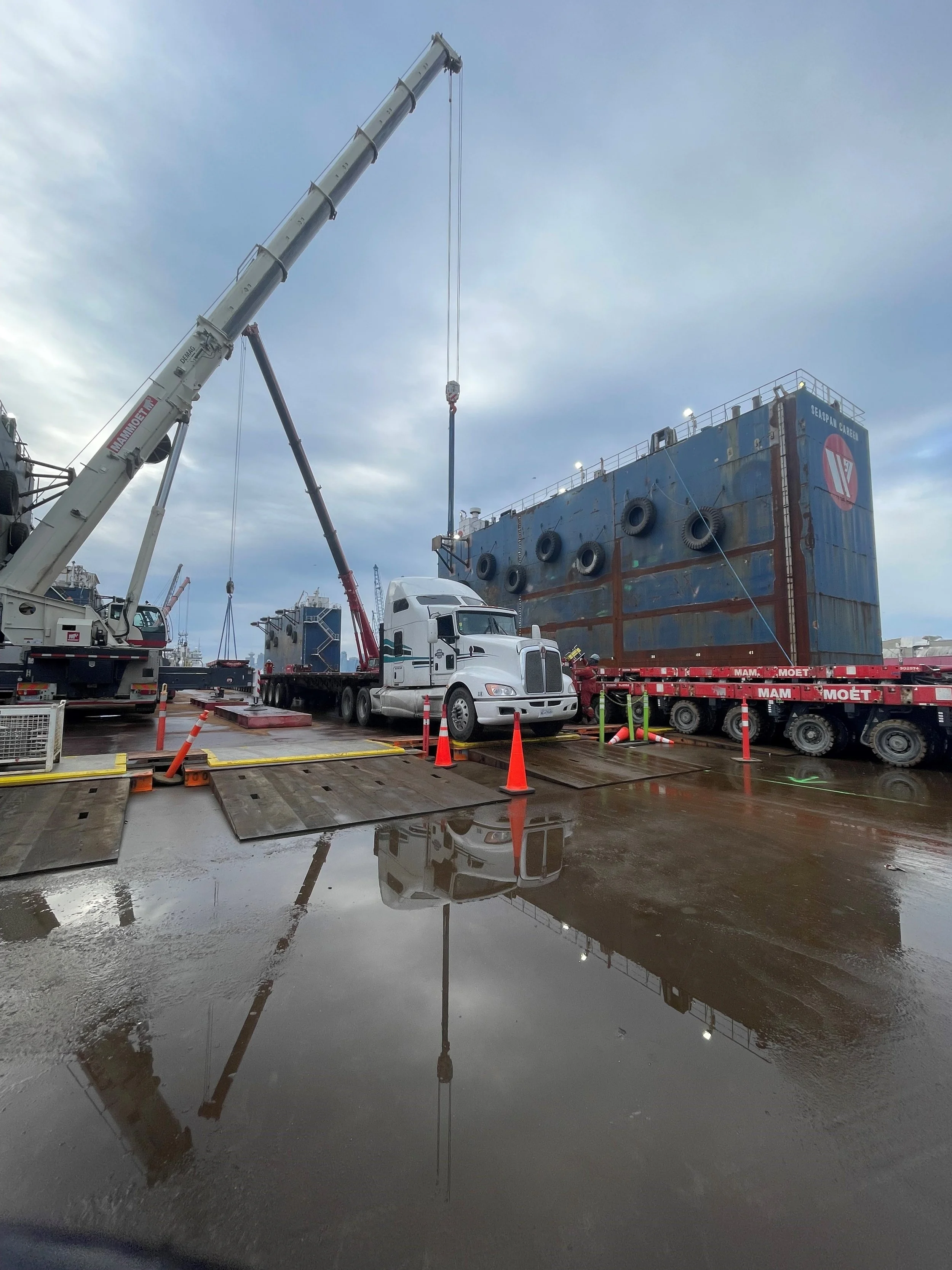 A crane unloading a large ship in a harbor, with wet ground and puddles reflecting the crane and ship.