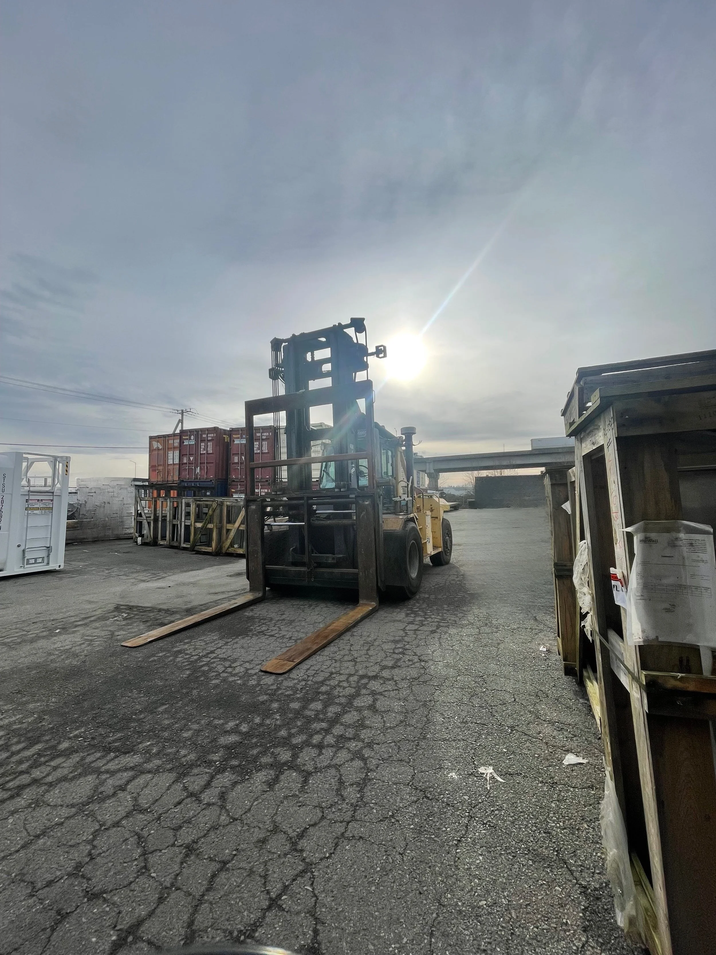A forklift on a cracked asphalt lot with stacks of shipping containers and a clear sky with the sun shining behind the forklift.