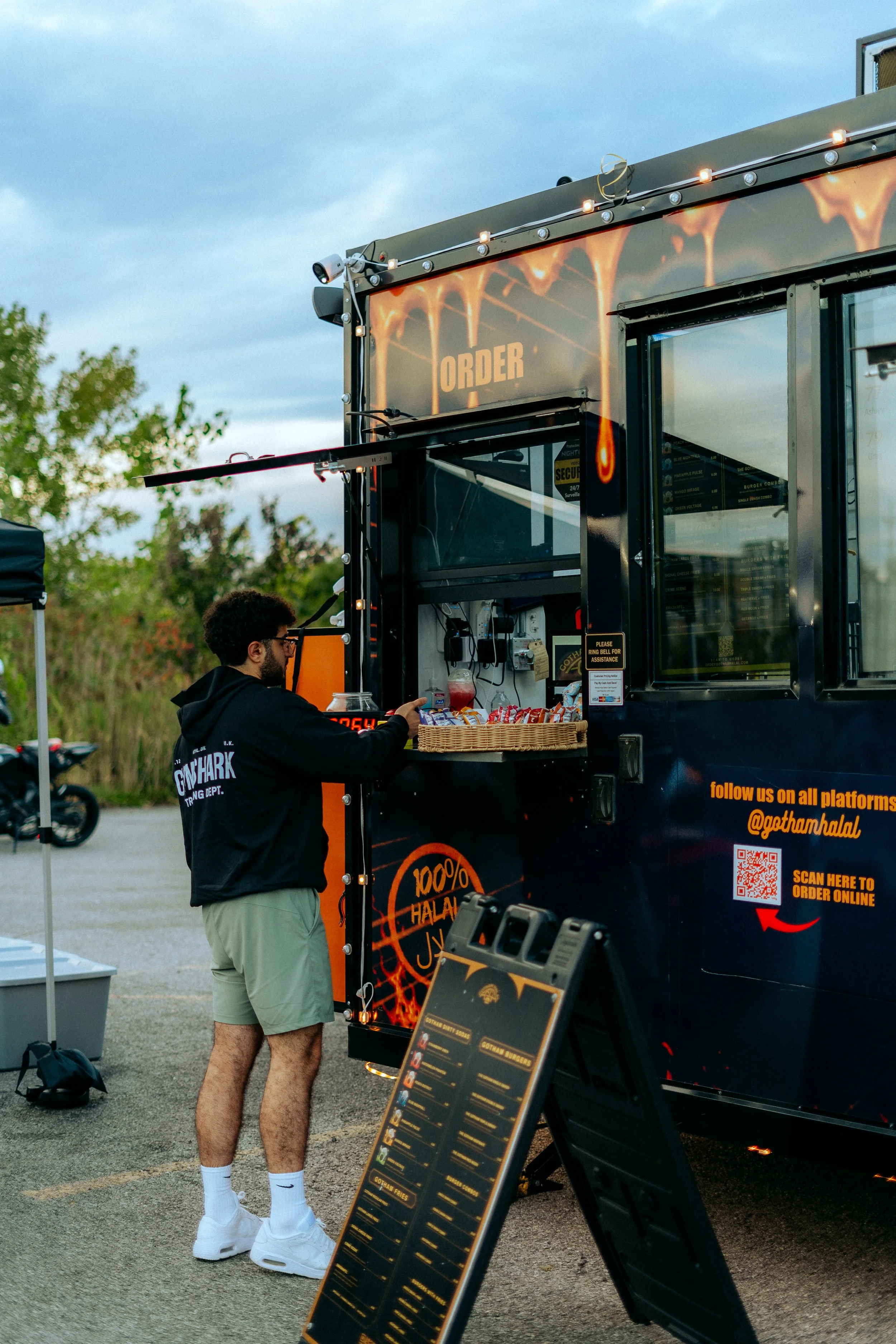 Person ordering food at a black food truck with a menu sign in the foreground, outdoors during daytime.