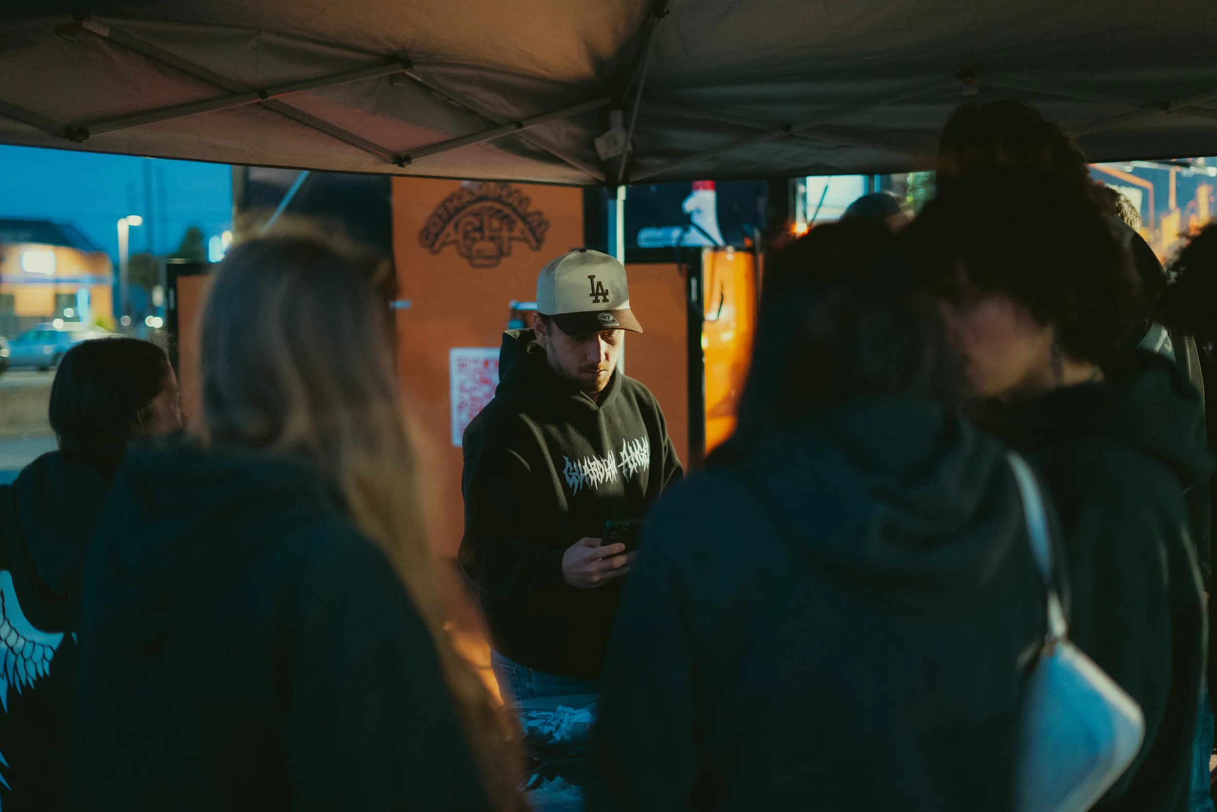 People gathered outdoors under a canopy at night, with one young man wearing a white Los Angeles Dodgers cap in the center looking at his phone, and surrounding individuals engaged in conversation or looking elsewhere.