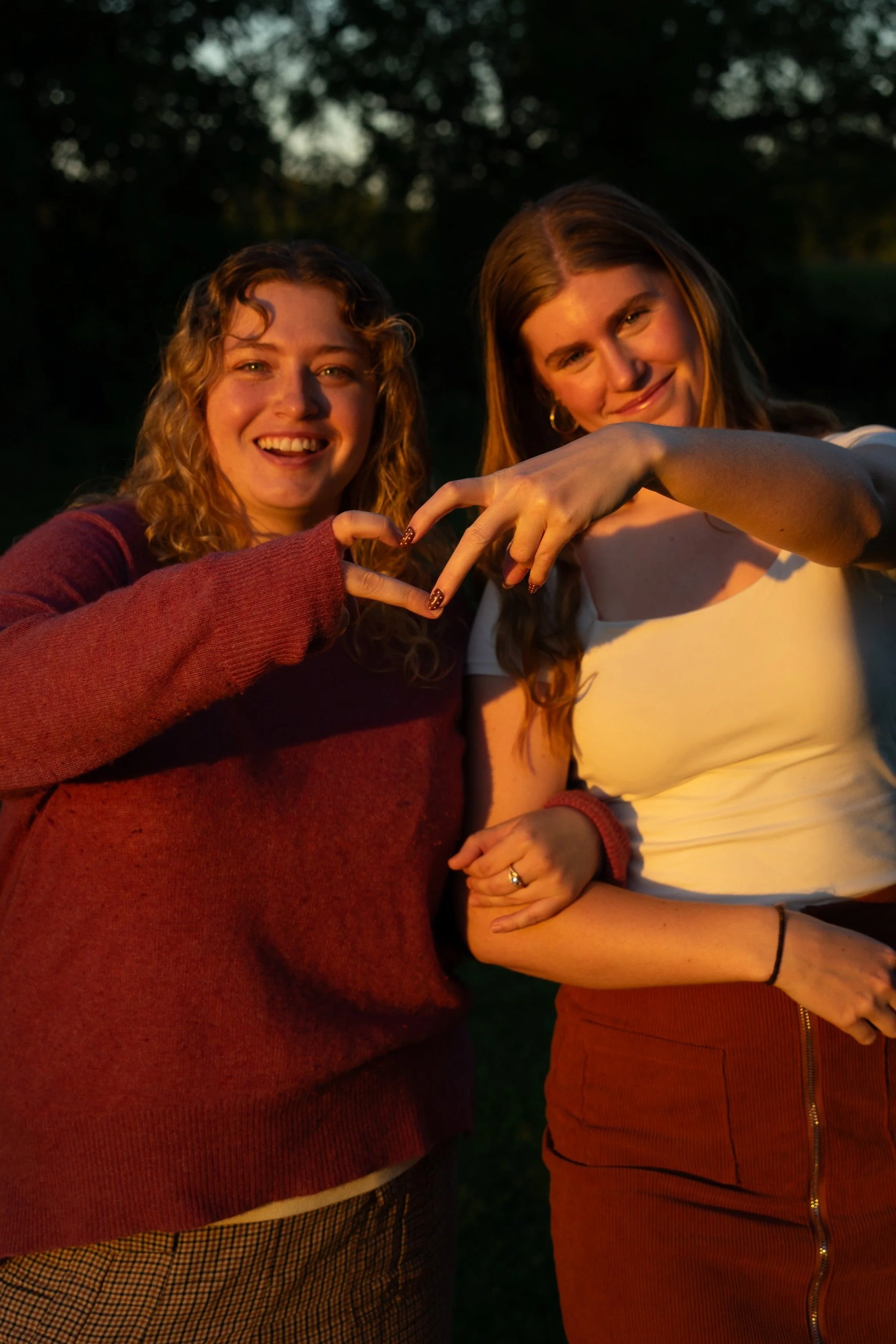 Two young women smiling and making a heart shape with their hands outdoors during sunset.