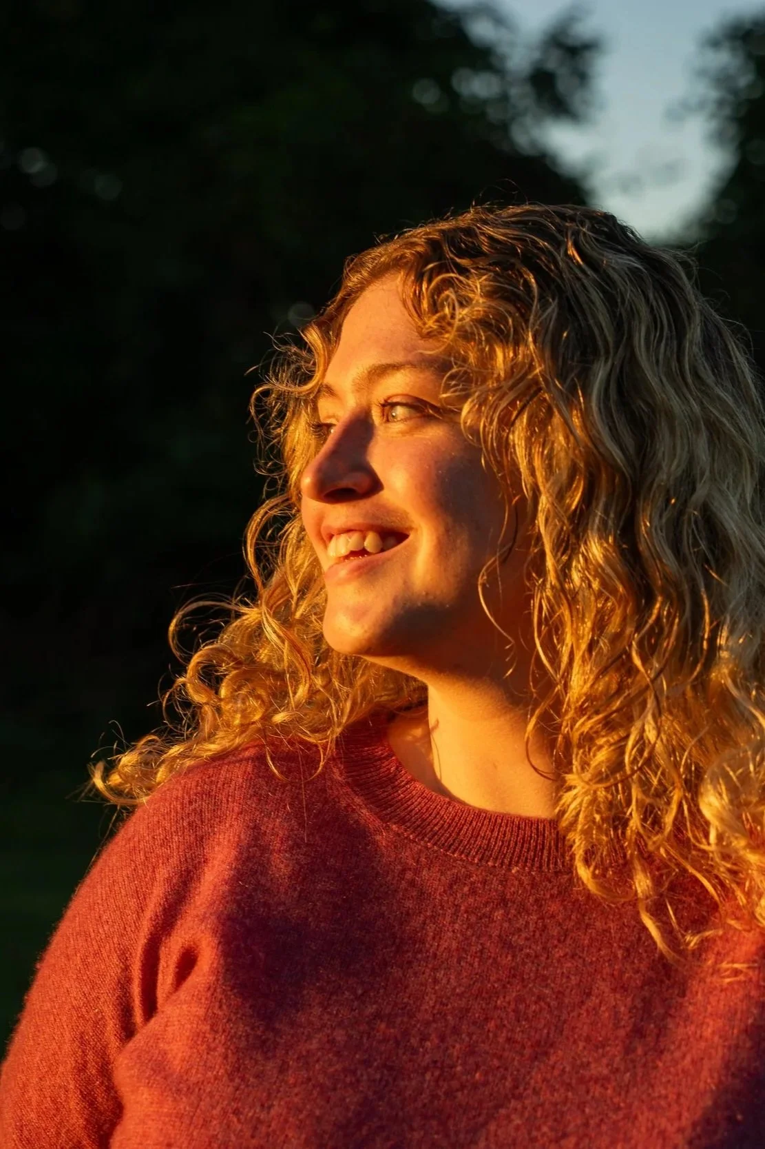 A woman with curly blonde hair smiling and looking to the side during sunset.