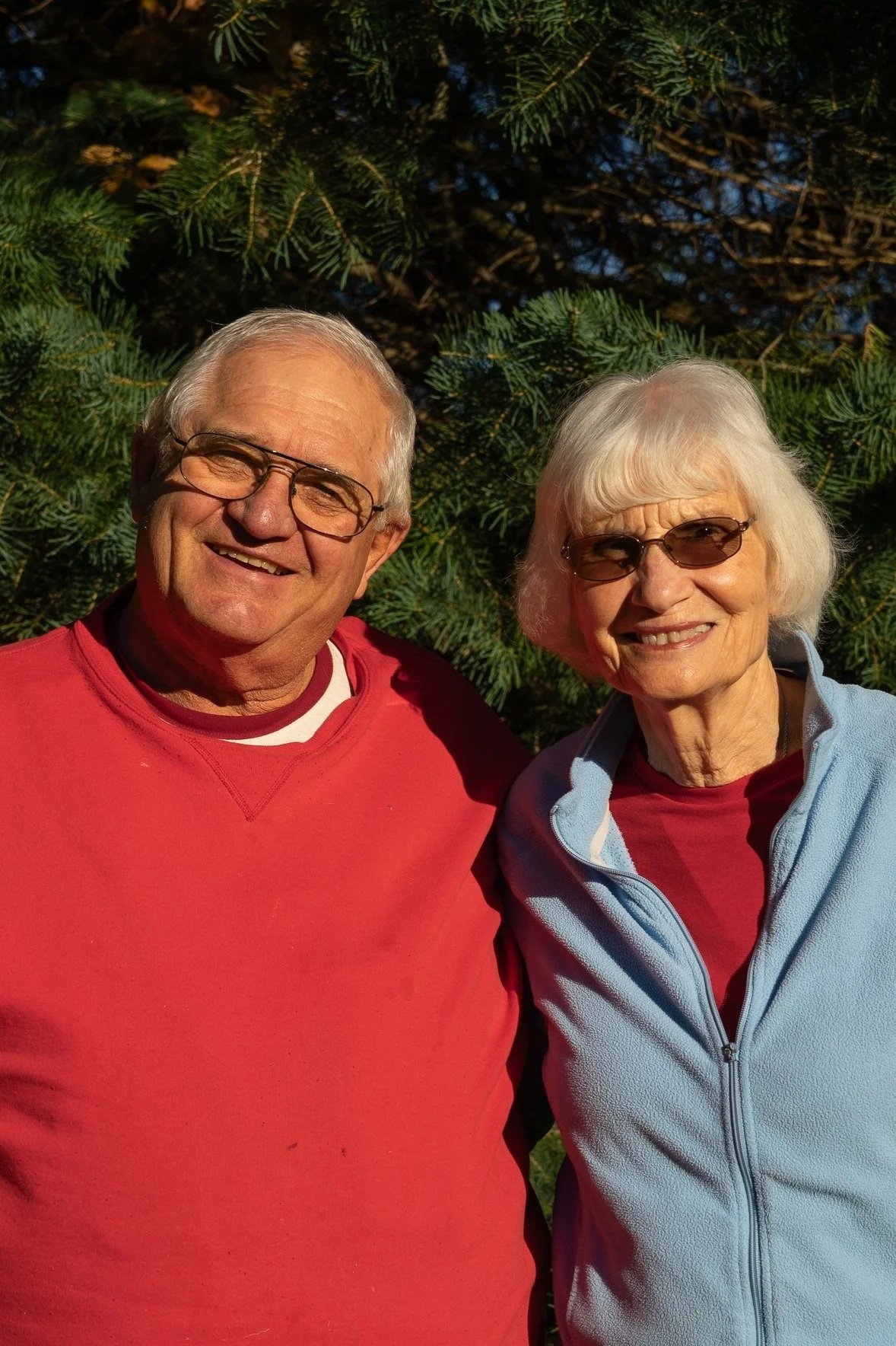 An elderly man and woman smiling outdoors in front of green foliage, both wearing sunglasses and casual clothing.