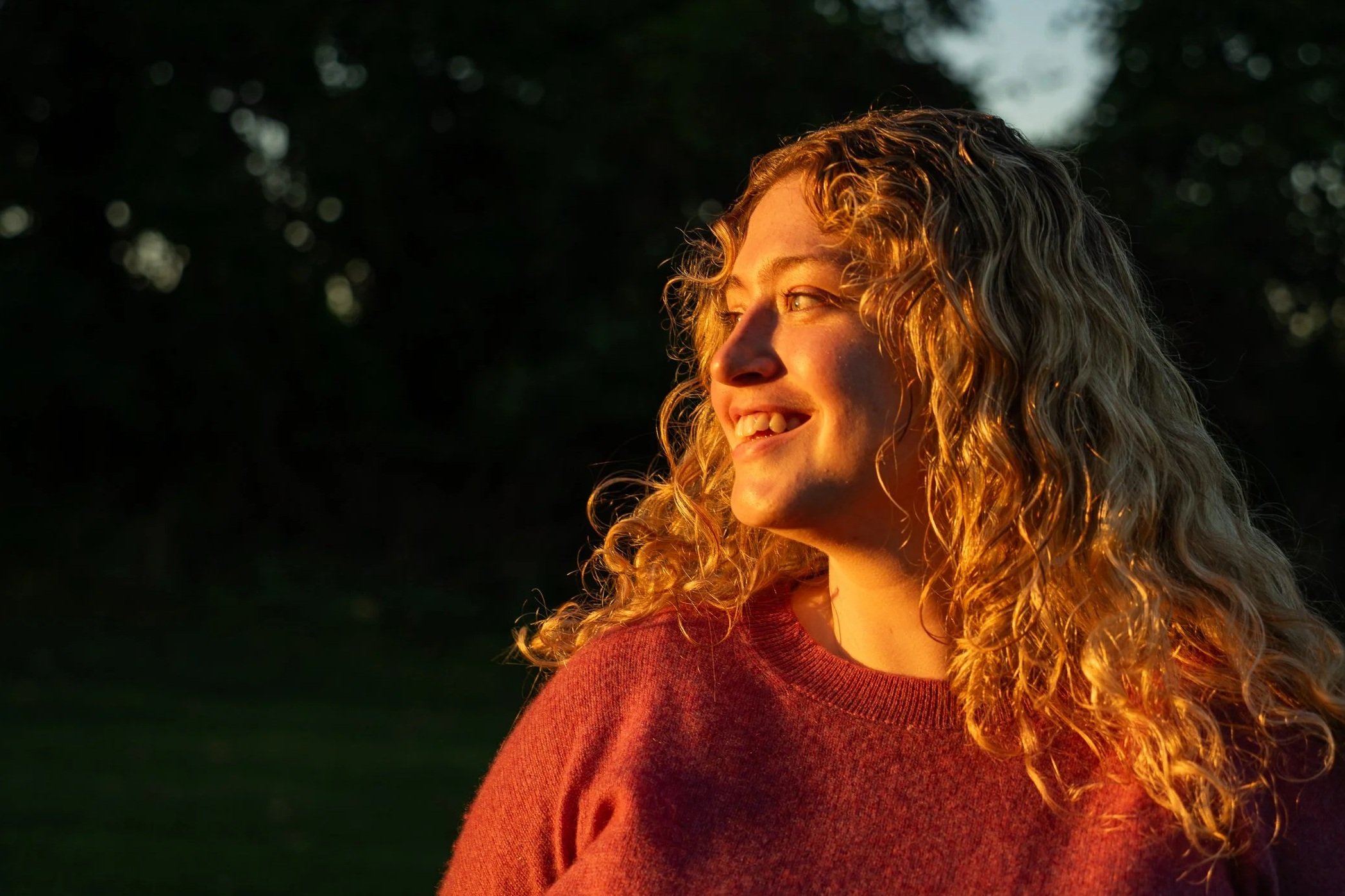 A woman with curly blonde hair smiling outdoors during sunset, wearing a red sweater.