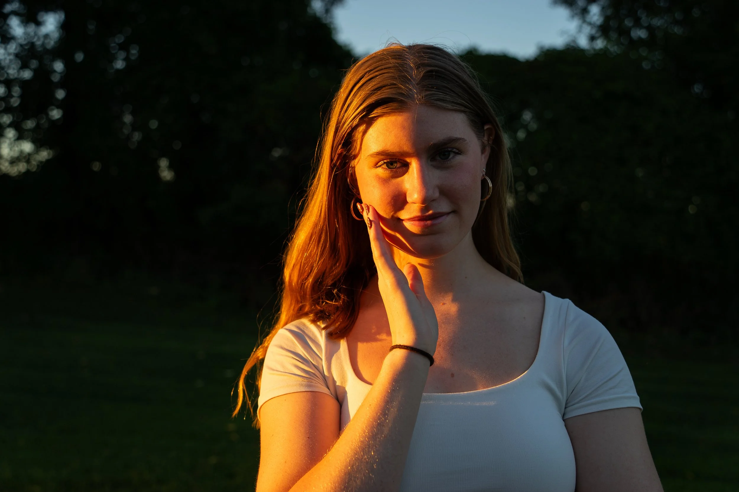 A young woman with long hair and hoop earrings is outdoors during sunset, touching her face with her right hand and looking at the camera. The setting sun casts a warm glow on her face and arm, with a blurred background of trees and sky.