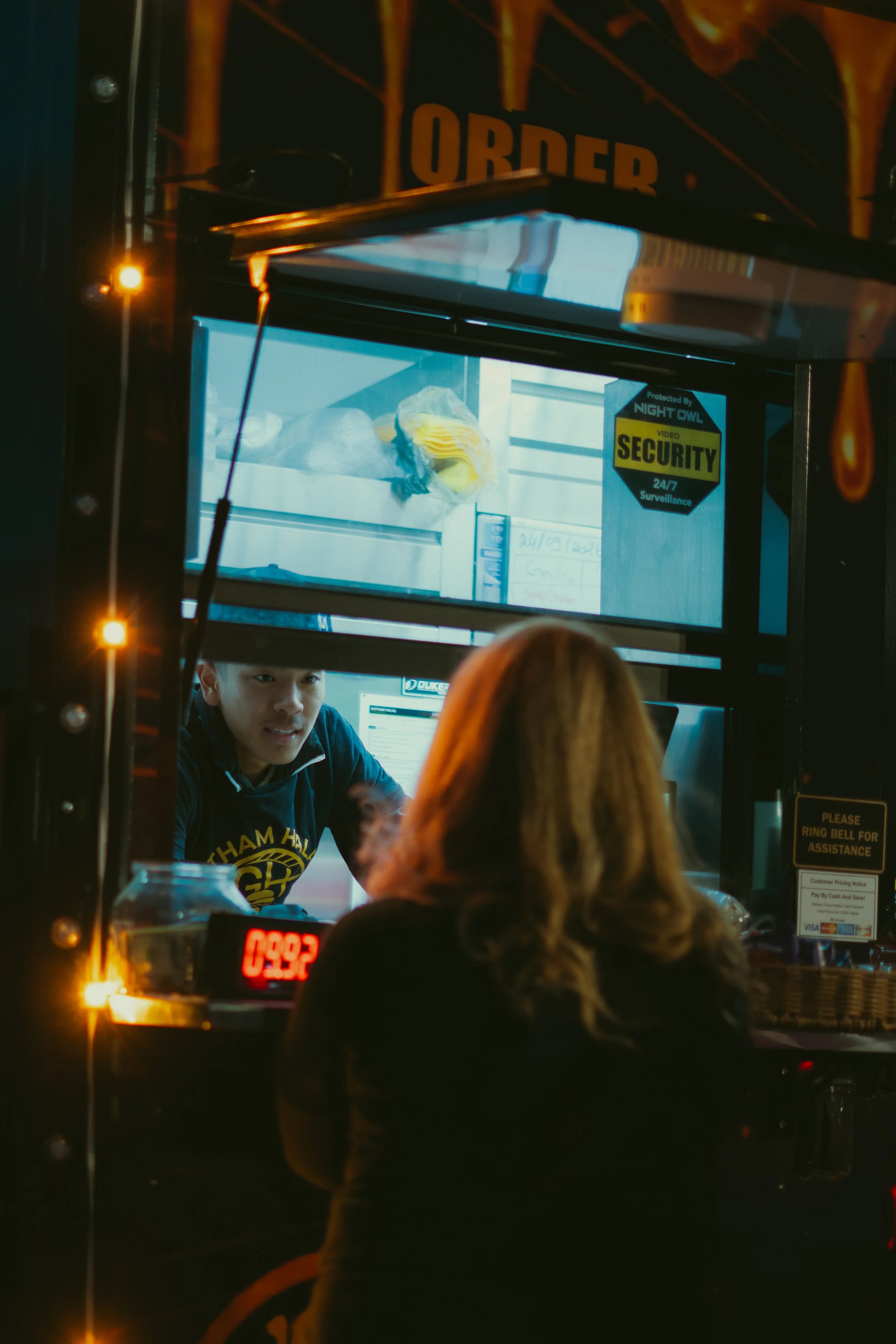 A food truck with a man inside, wearing a Golden State Warriors hoodie, talking to a customer with long hair. The truck has a digital order display and security signage, illuminated by warm lighting.