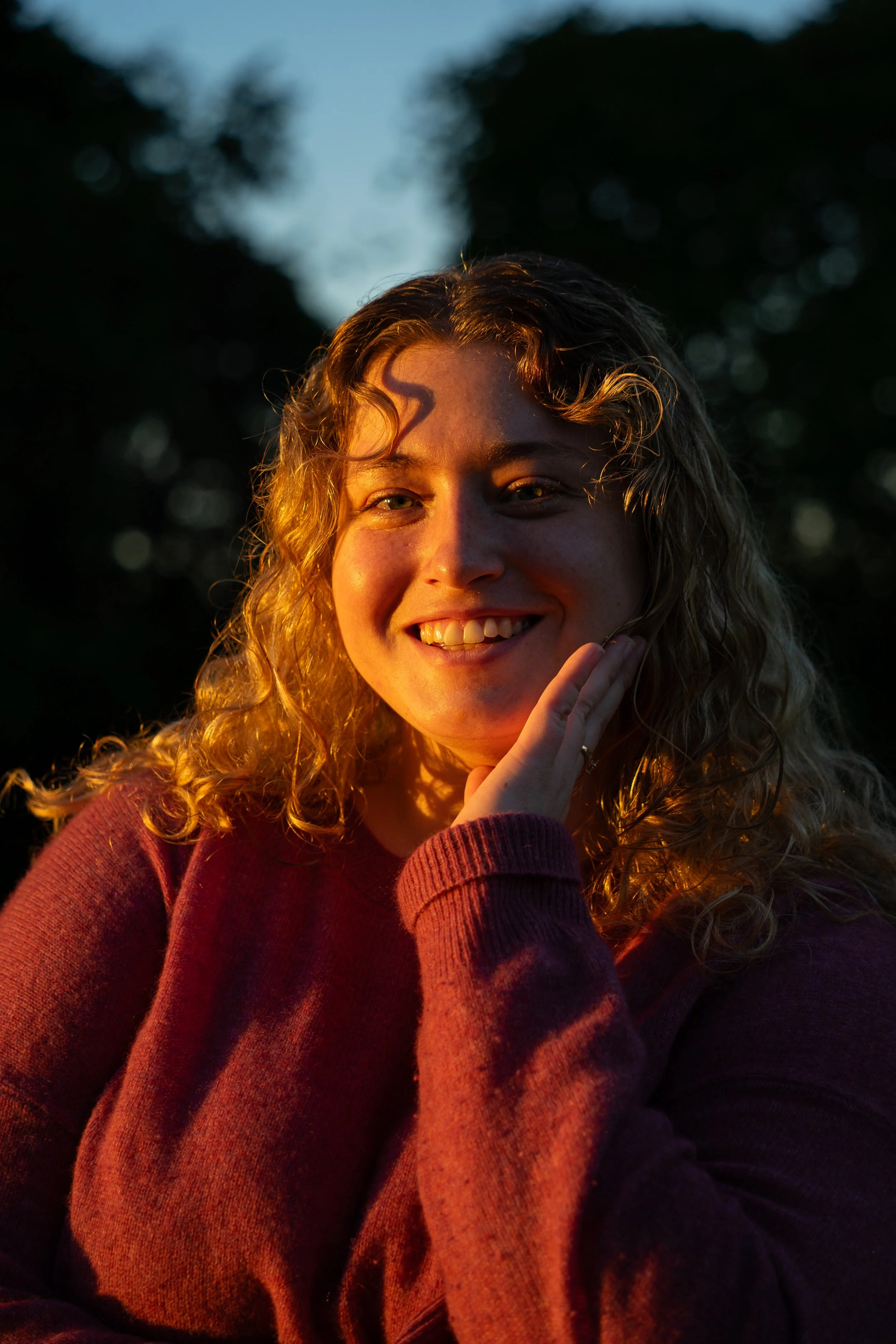A young woman with curly blonde hair smiling, wearing a maroon sweater, outdoors during golden hour with dark trees in the background.