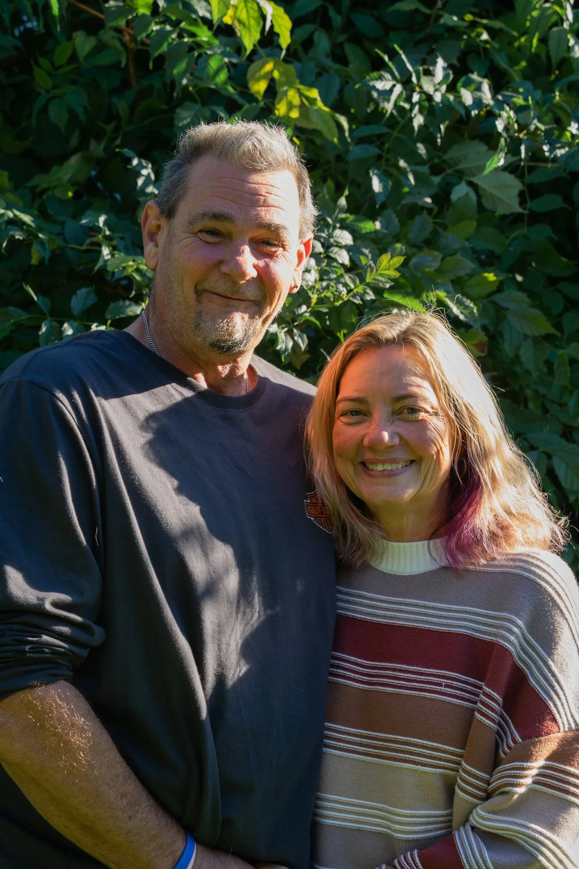 A man and a woman standing outdoors in front of green foliage, smiling at the camera, with the man wearing a black shirt and the woman wearing a striped sweater.
