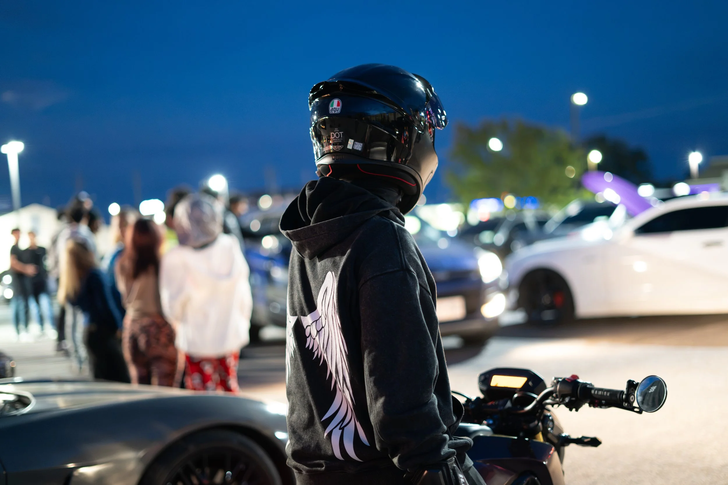 Person wearing a black helmet and hoodie, standing next to a motorcycle at night in a parking lot filled with people and cars.