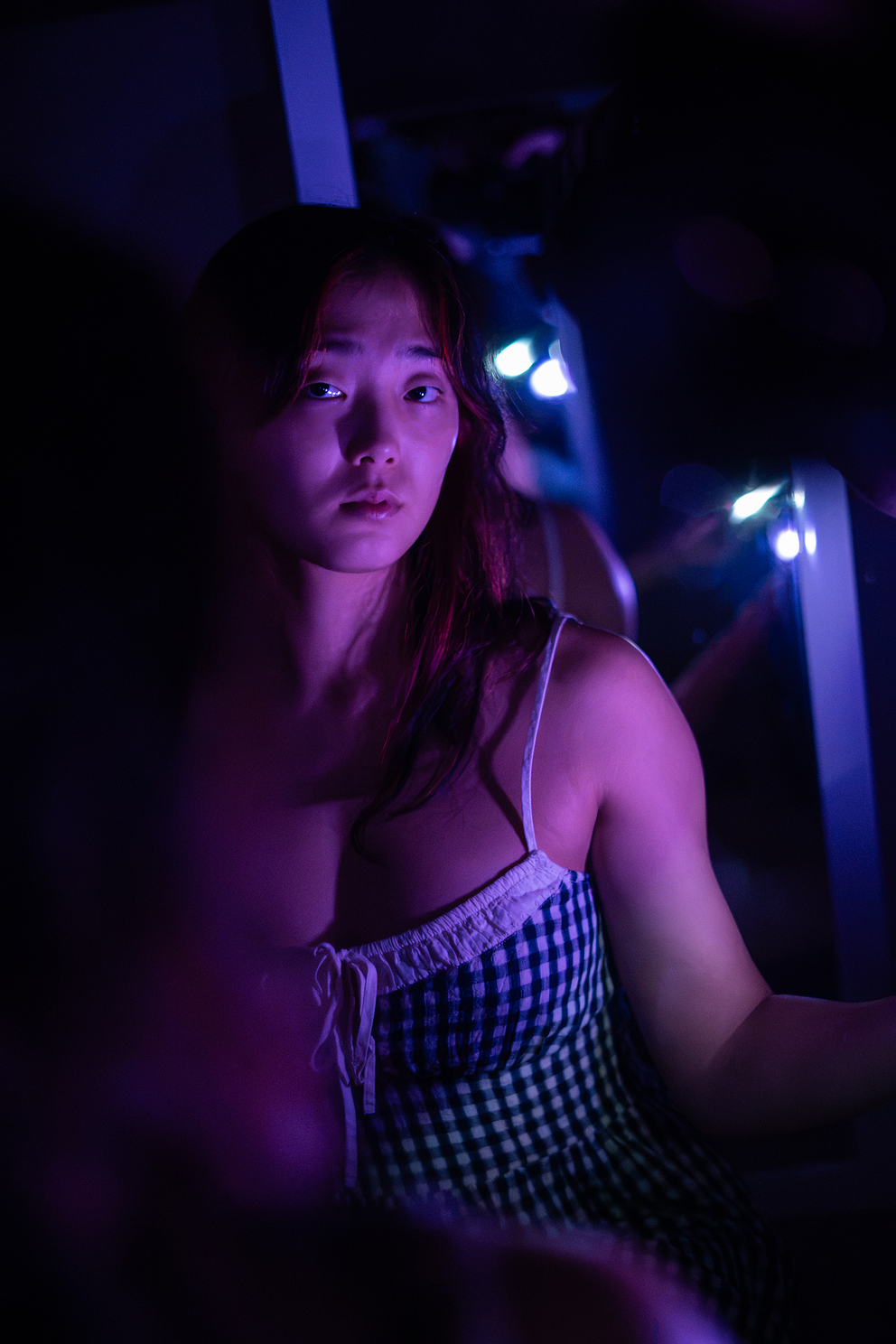 A young woman with long hair, wearing a checkered dress, is sitting in a dimly lit room with purple and blue lighting, looking at the camera.