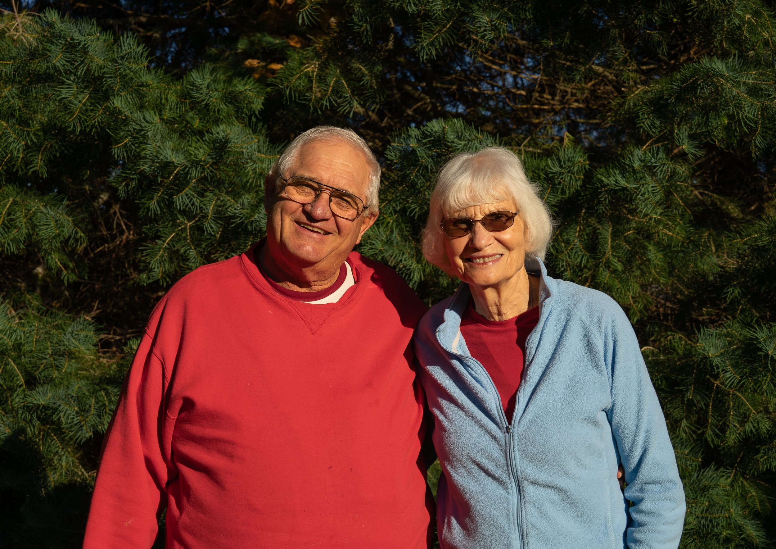 An elderly couple smiling outdoors in front of green pine trees.