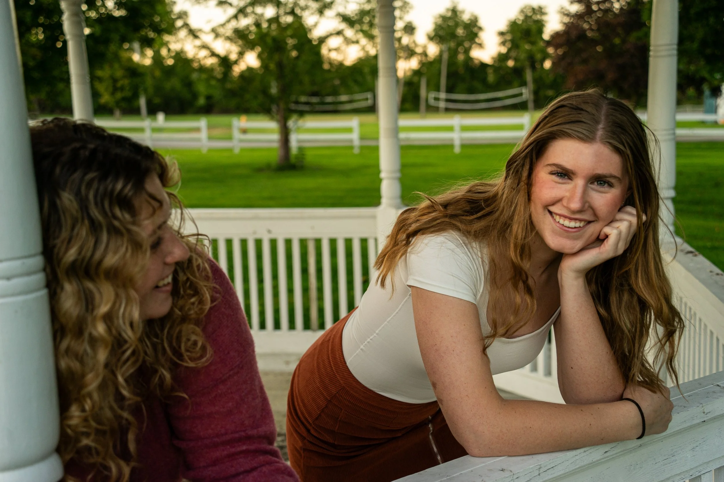 Two young women with red hair sitting on a porch, smiling and engaging in conversation. One is wearing a white shirt and rust-colored pants, and the other is wearing a maroon sweater. The background includes green trees and a white fence.