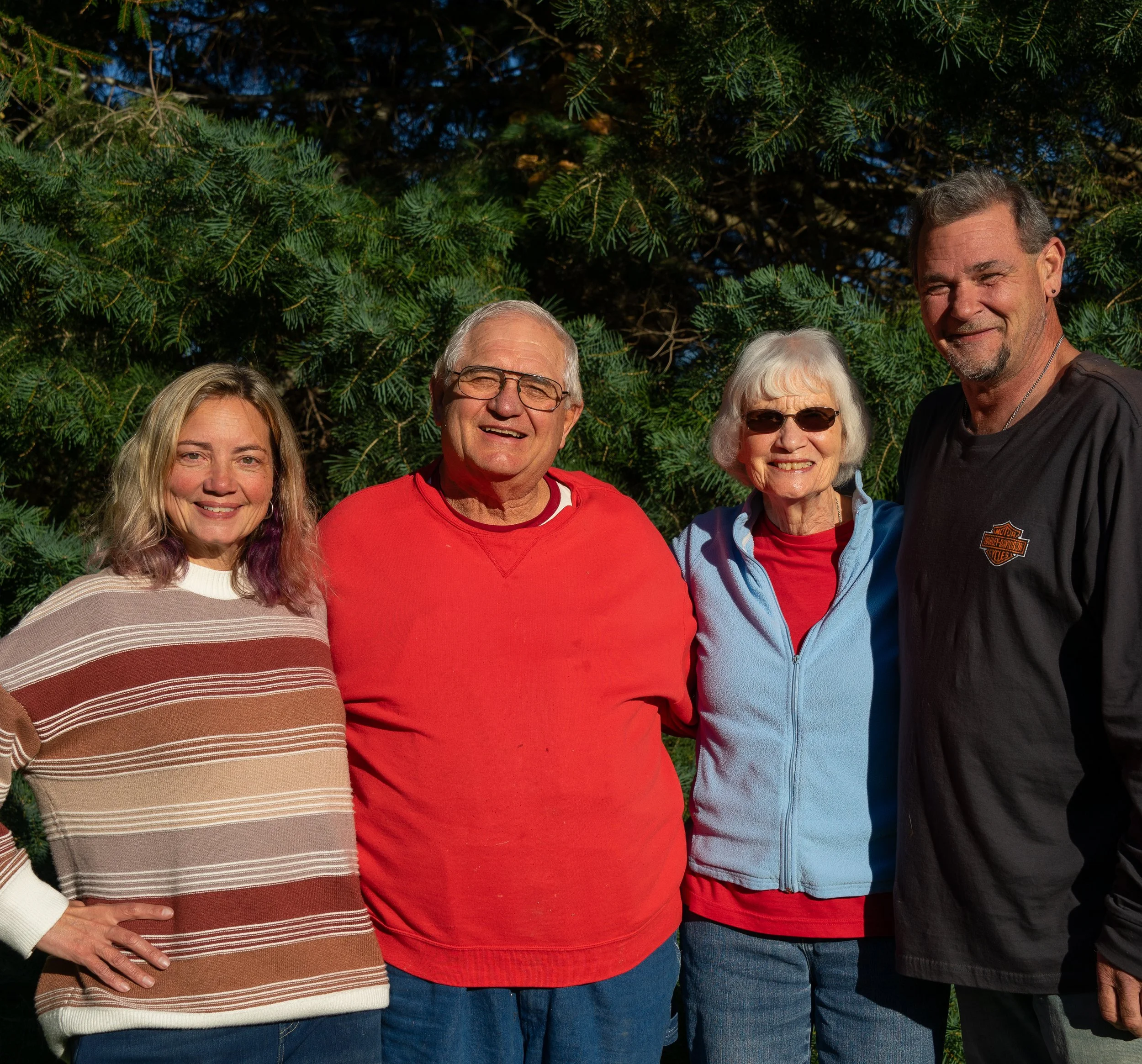 An elderly couple, their daughter, and their son-in-law are standing outdoors in front of a pine tree, smiling and wearing casual clothing, with some wearing sunglasses.