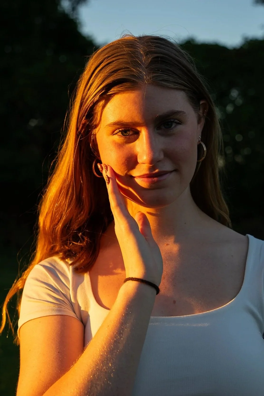 A young woman with long hair, wearing a white shirt, gently touching her face with her right hand. She is outdoors at sunset, with warm sunlight illuminating one side of her face and hair, and a dark, blurred background of trees.