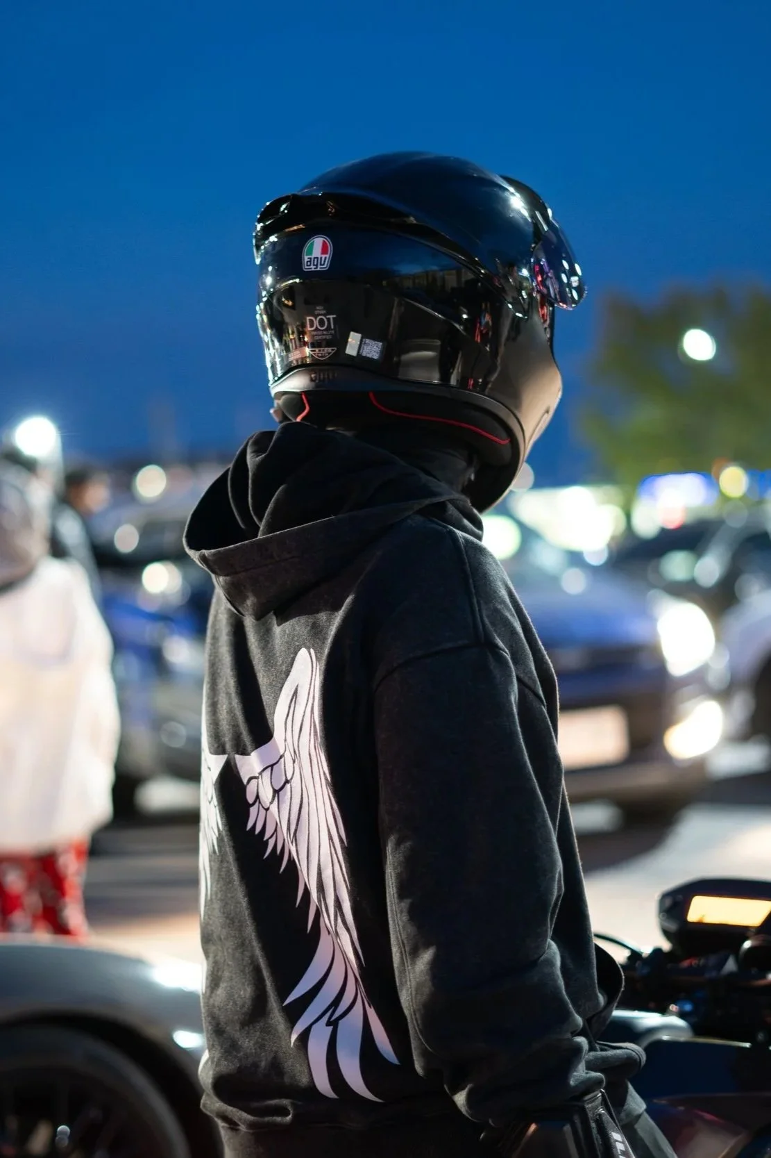 Person wearing a black helmet and black hoodie with a white wing design, standing in a parking lot during dusk or night with blurred cars and people in the background.