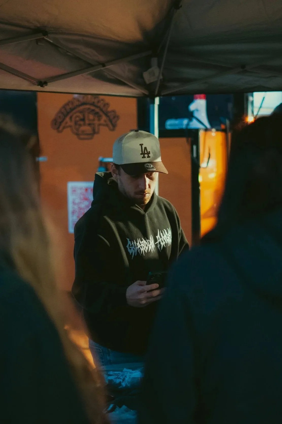 A man in a black hoodie with white text and a beige LA baseball cap is looking at his phone. There are several people around him, and the setting appears to be a night outdoor event, possibly a food stand or booth, with a sign and a QR code on the wall behind him.
