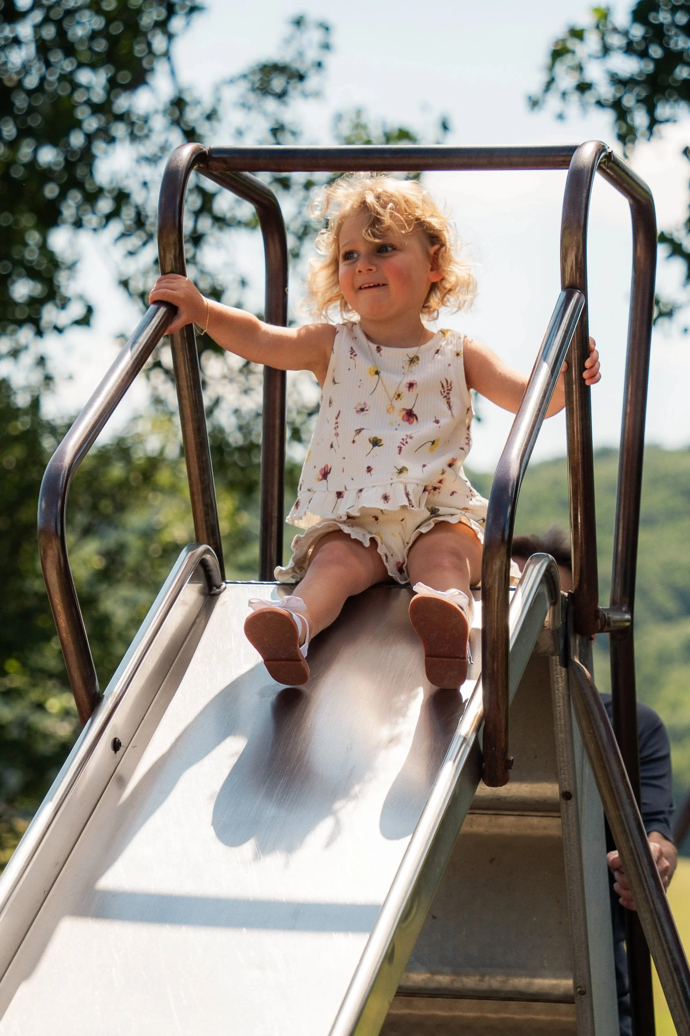 A young girl with curly blonde hair and wearing a floral dress is sitting at the top of a metal slide at a playground, holding the sides of the slide, with trees and a cloudy sky in the background.
