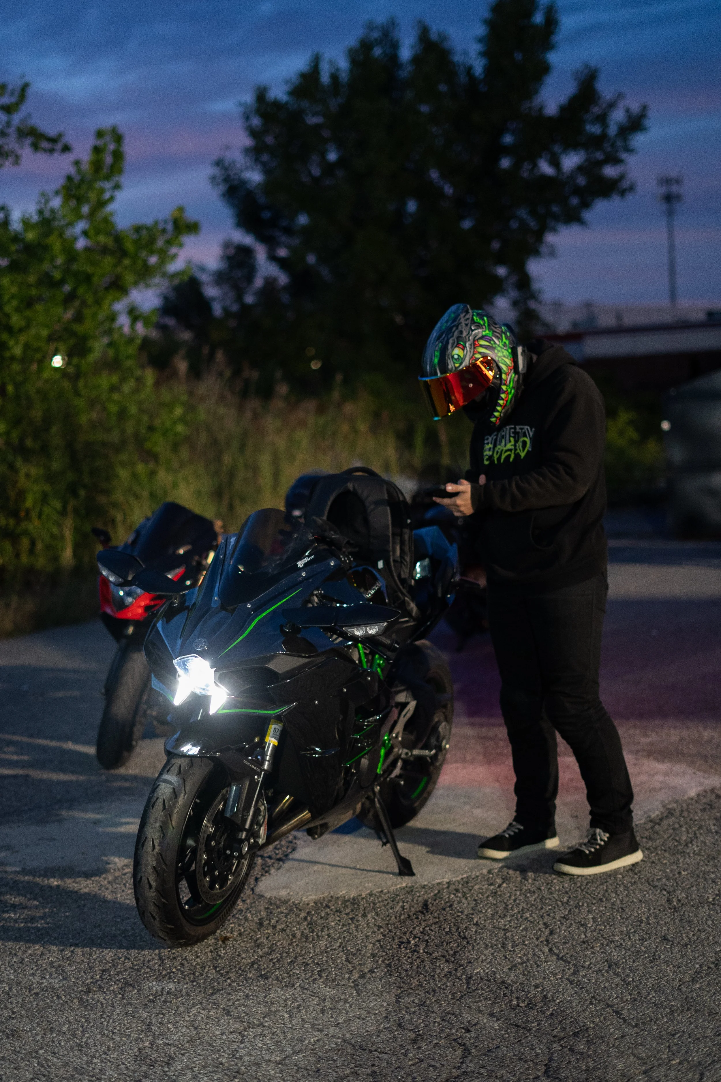 A person wearing a colorful, monster-themed helmet and black hoodie stands next to a black motorcycle with green accents during dusk or early evening. Trees and parked cars are visible in the background.