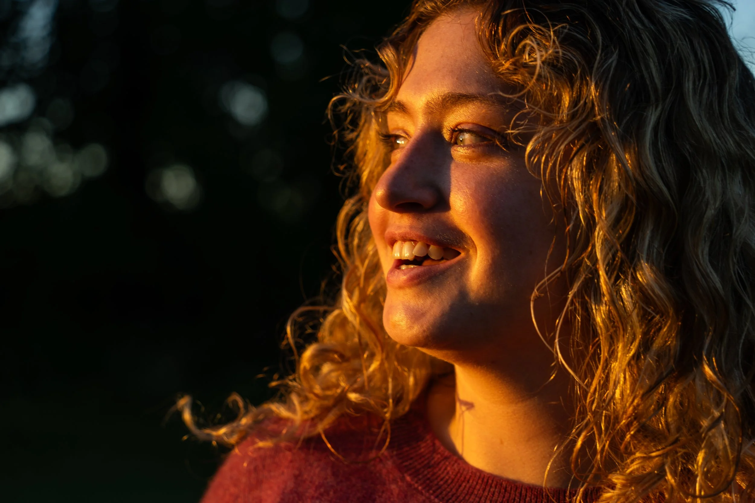 Close-up of a young woman with curly blonde hair smiling as she looks towards the sunlight, with warm lighting highlighting her features.