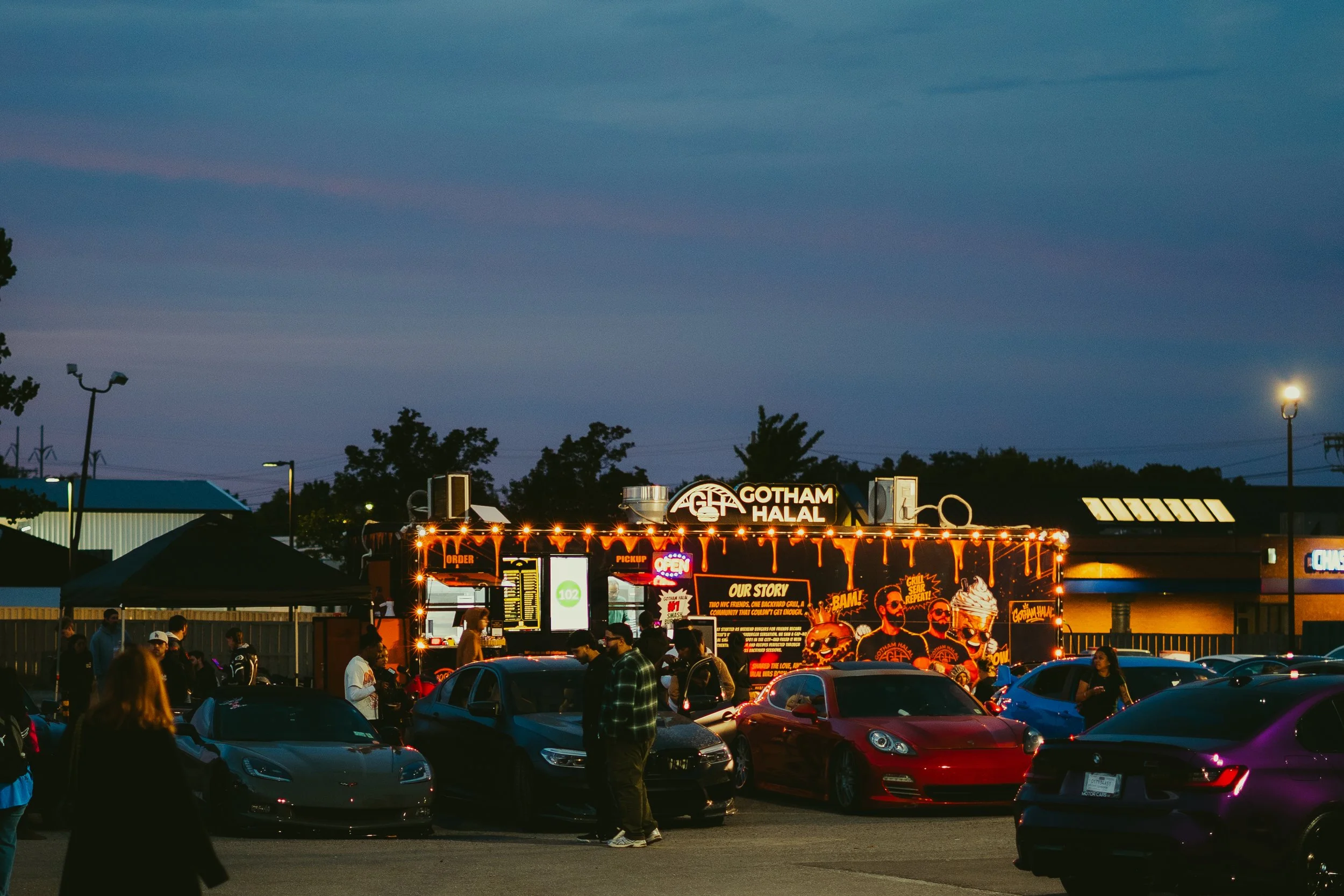 Parking lot filled with cars and people, food truck with illuminated signs and Gotham Halal branding, trees and commercial buildings in the background, evening sky with clouds.