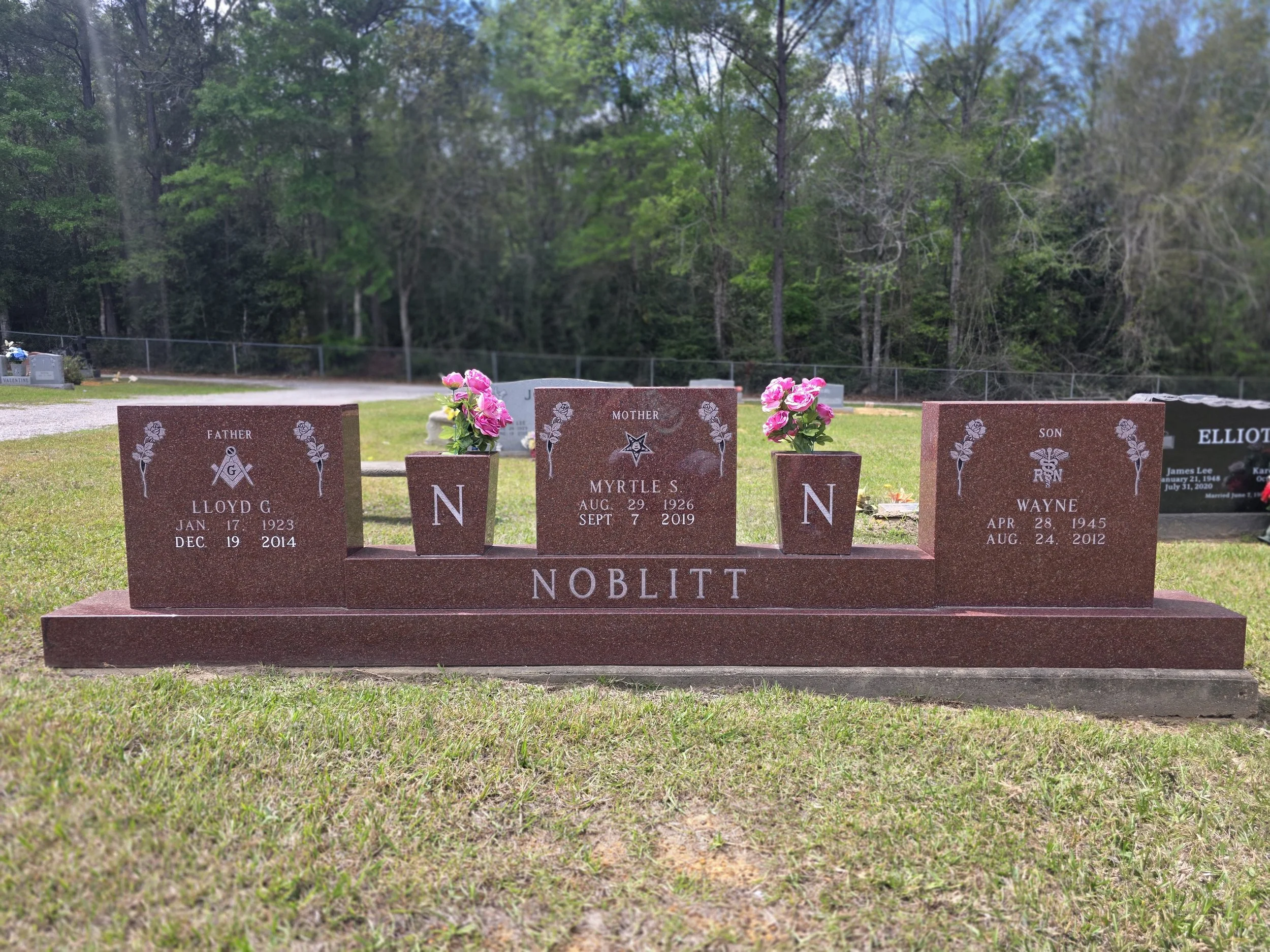 Red granite family tombstone with three sections, dedicated to Lloyd G. Noblitt, Myrtle S. Noblitt, and Wayne Noblitt, adorned with pink flowers in vases, set in a grassy cemetery with trees in the background.