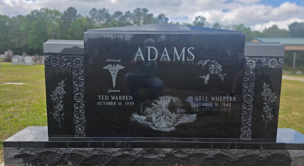 A black granite tombstone with the surname 'Adams' engraved at the top. The tombstone commemorates Ted Warren, born October 16, 1939, and Jo Nell Wheeler, born December 31, 1942. It features symbolic engravings of a medical caduceus, a dove with flowers, and a garden gate with a cross and trees, set in a cemetery with other graves visible in the background.