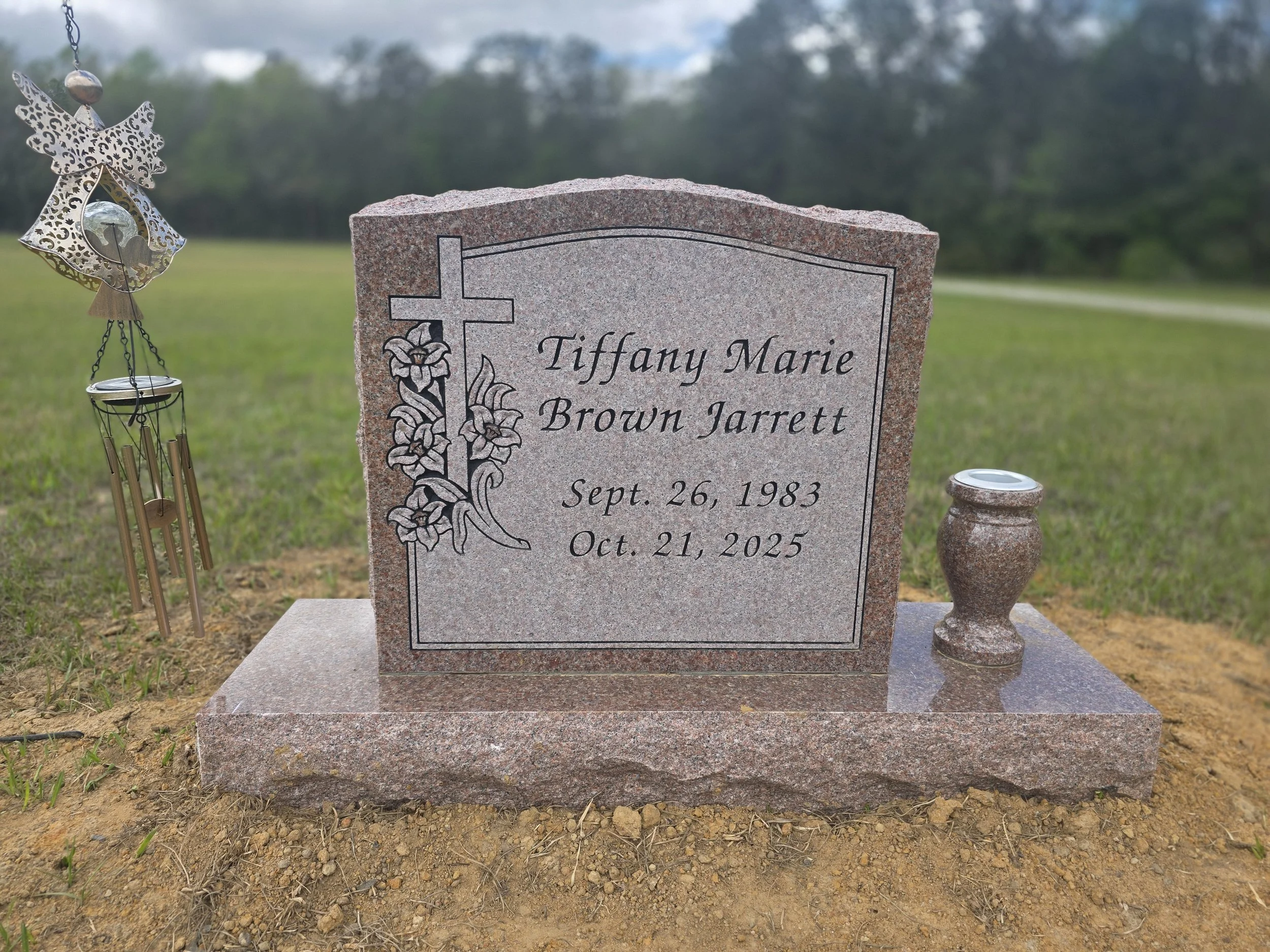Gravestone for Tiffany Marie Brown Jarrett, with a cross and floral design, set in a grassy area, with a wind chime and candle holder nearby, against a background of trees and sky.