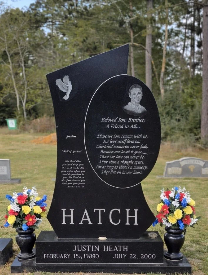 Black headstone  surrounded by flowers. The headstone has a dove at the top and an inscription that reads, “Beloved Son, Brother, A Friend to All...”