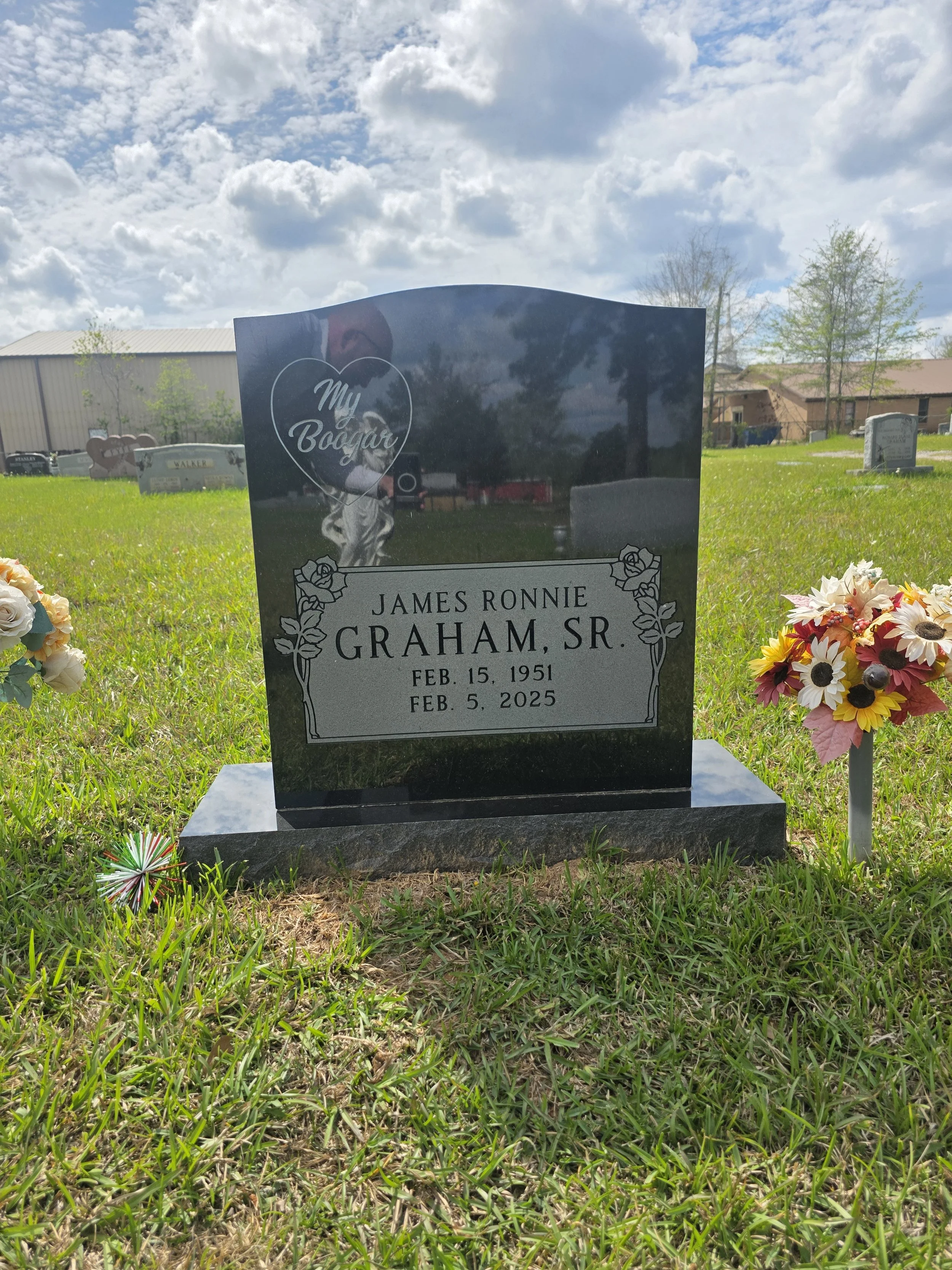Gravestone for James Ronnie Graham Sr., with the dates February 15, 1951 to February 5, 2025, in a cemetery with grass and flowers, under a partly cloudy sky.