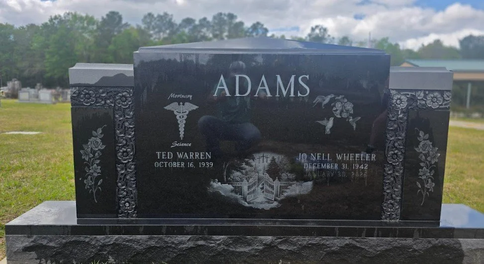 Black granite headstone for the Adams family with floral engravings and religious symbols, including a caduceus with a torch, and images of a dove, flowers, and a gated garden, engraved with names and dates of Ted Warren and Jo Nell Wheeler.