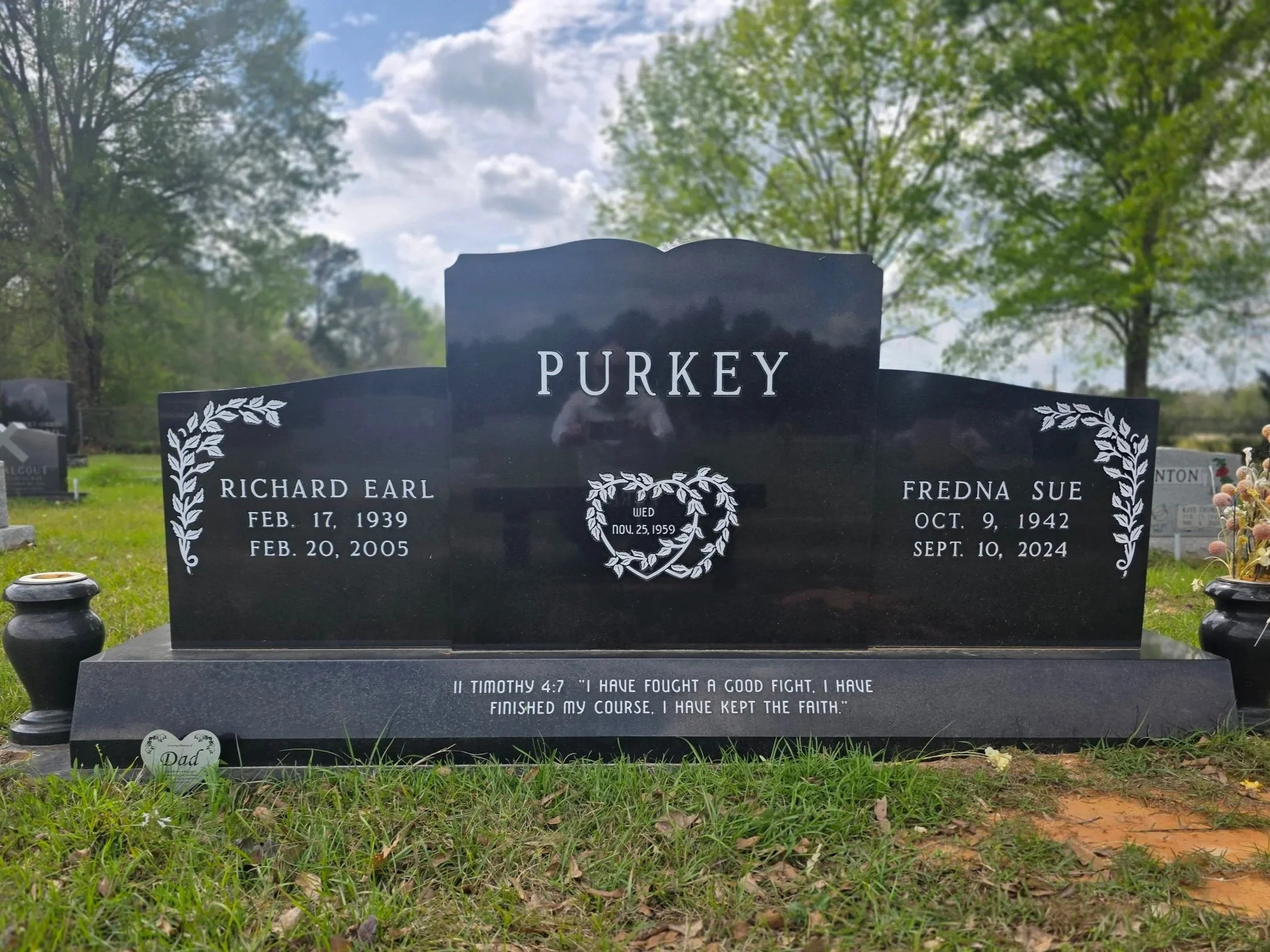 A black granite headstone at a cemetery.  An inspirational quote from 1 Timothy 4:7 is inscribed at the bottom. Small vases with flowers are positioned on either side of the headstone.