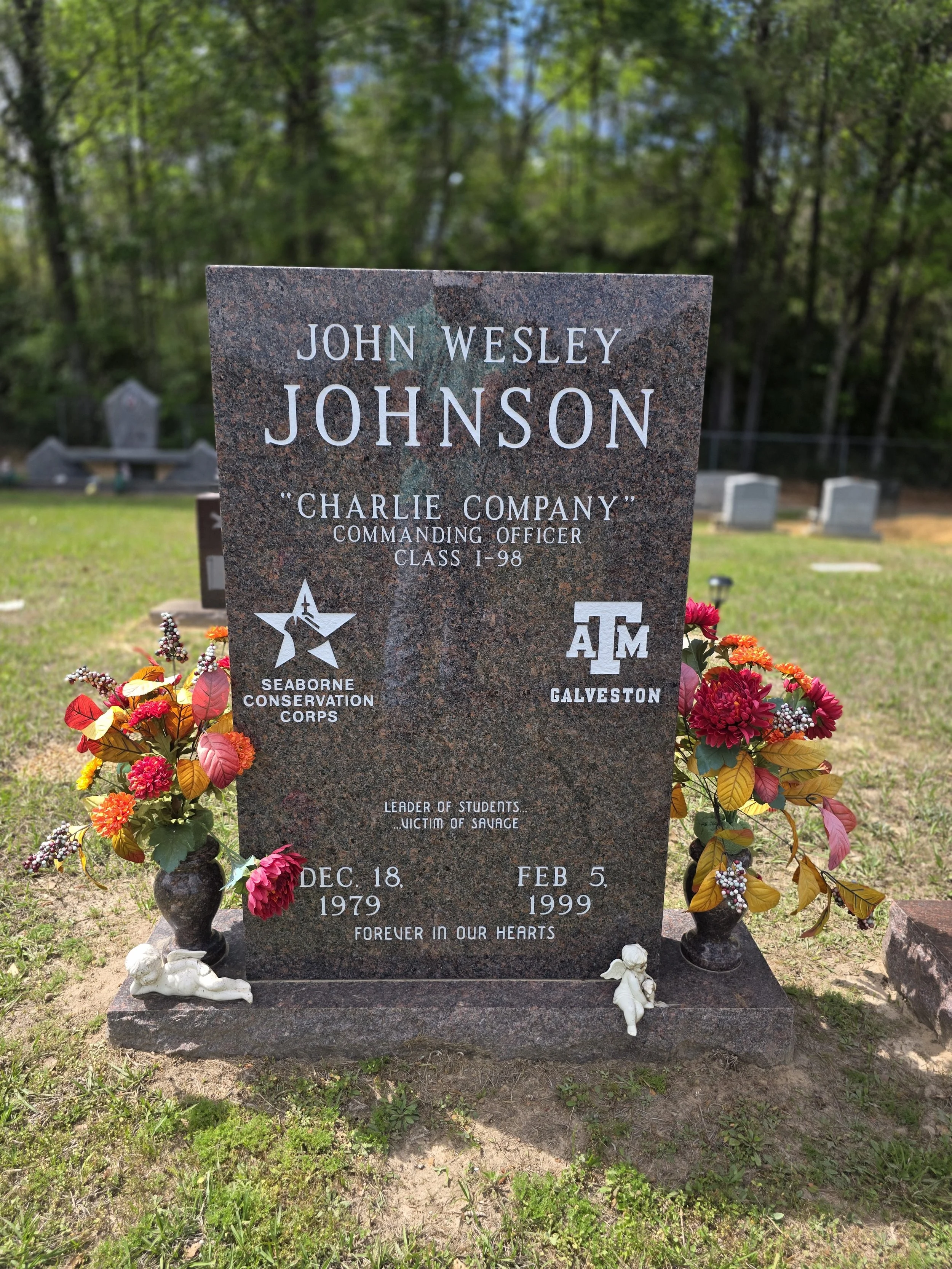 Gravestone with flowers and angel figurines. It reads 'John Wesley Johnson,' born December 18, 1979, died February 5, 1999, addressing him as a leader of students and victim of savage, with a tribute of forever in our hearts.